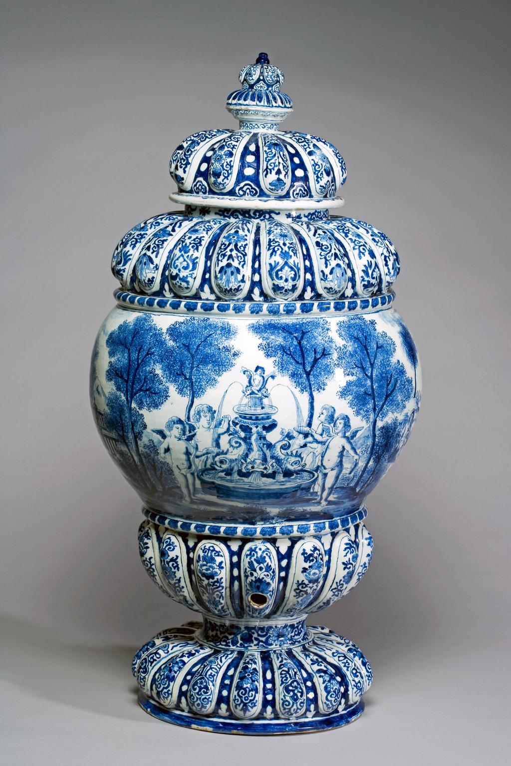 An image of Delftware. Cistern. Unknown Delft factory, Holland. The gadroons are decorated with Chinese-style flowers and symmetrical scrolls of two alternating designs. The convex area is painted with a continuous European landscape with buildings and sponged trees and bushes, amongst which are several men, one of them on a horse, and another smoking a pipe. To one side is a garden with a balustrade and a fountain with dolphins, round which stand four putti. Pale buff earthenware thrown, with applied moulded gadrooning, tin-glazed, and painted in blue, circa 1700. Baroque.