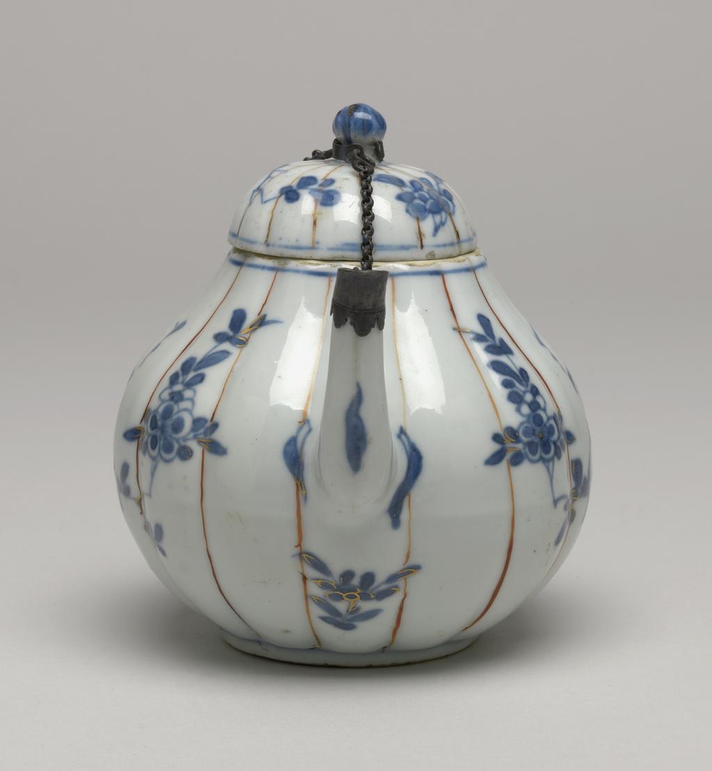 An image of Teapot and cover. Unknown production, Japan & China. The squat, pear-shaped body divided into fourteen lobes separated by single vertical lines in iron-red and painted on either side with a scene of a song-bird perched in the branches of a flowering prunus tree, in underglaze blue with details picked out in gilding The loop handle and upright spout are attached by a detachable metal chain to the globular knop at the centre of the cover, thus holding it in place on the teapot. The cover is lobed to match the teapot and decorated with two sprays of peony in underglaze blue. Hard-paste porcelain painted underglaze in blue with iron-red enamel, and gilding, height, whole, 11 cm, diameter, whole, 16.2 cm, circa 1662-circa 1722. Chinese Imari. Qing Dynasty (1644-1912). Kangxi Period (1662-1722).