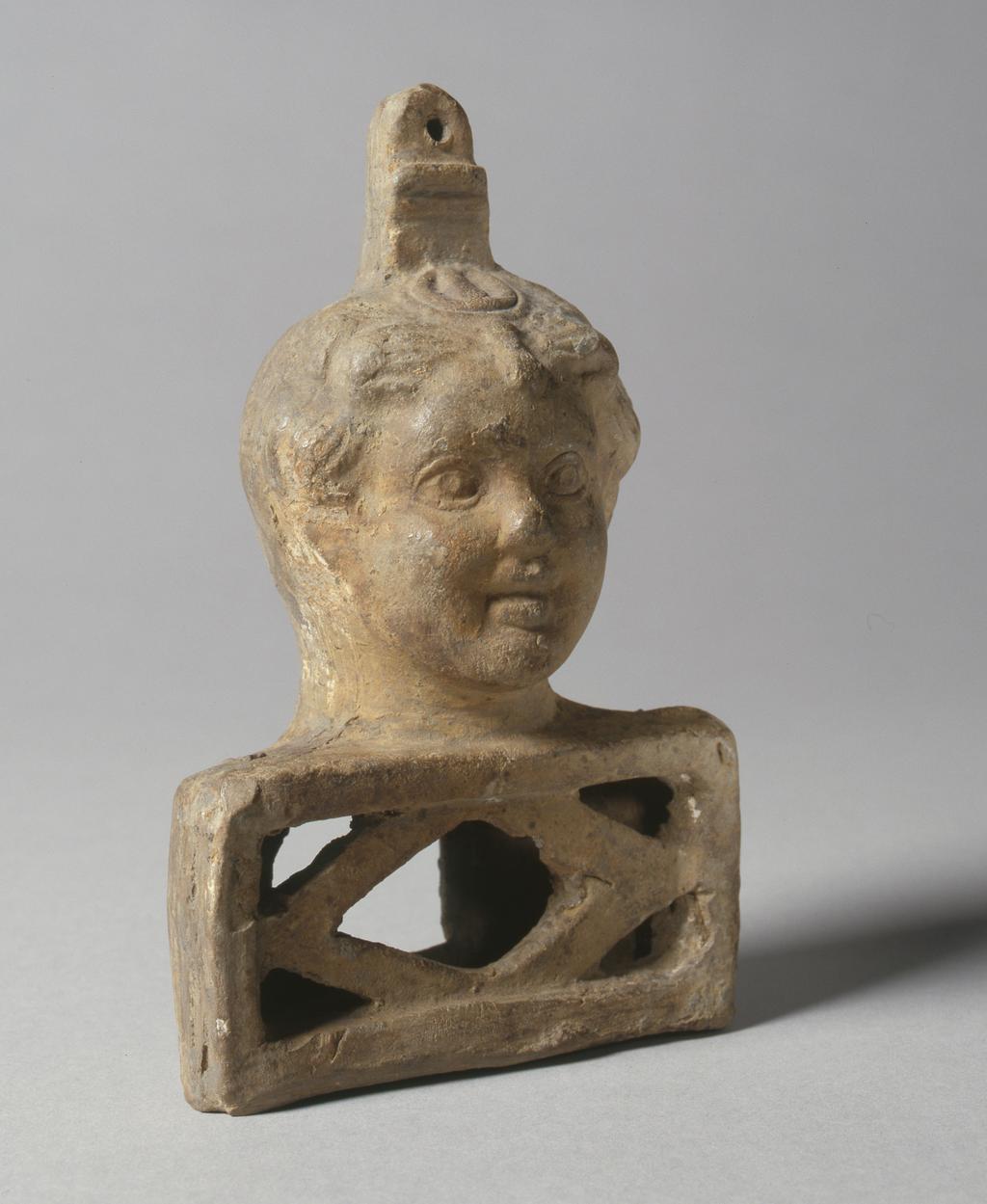 An image of Equipment/utensil. Clay lamp holder in the form of Harpocrates. With youthful male head on rectangular plinth - a small votive lamp would have been placed in the bottom section. Find Spot/Production Place: Egypt. Clay, mould-made, length 14.75 cm, circa A.D.100-200.