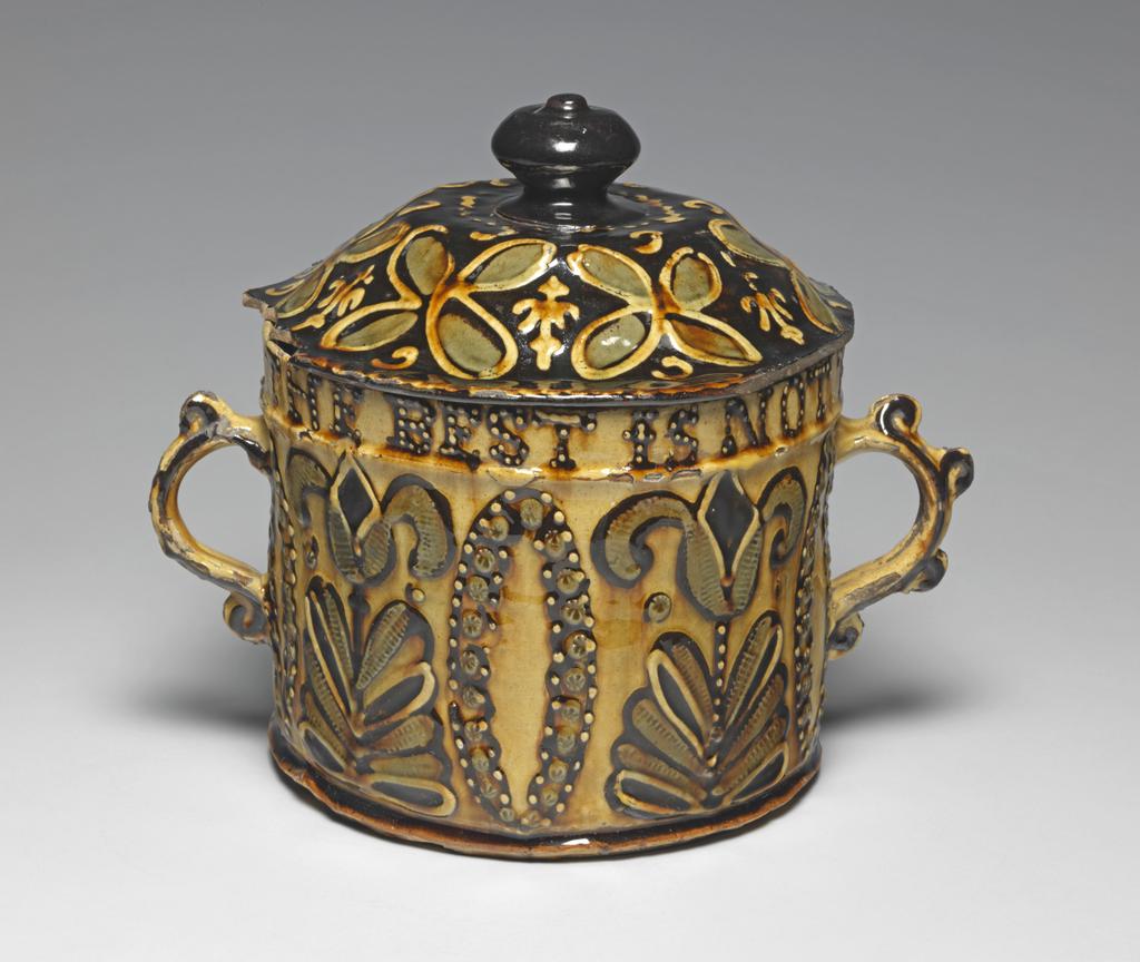 An image of Posset Pot/Two-handled pot and cover. Staffordshire Pottery, unknown maker. Red earthenware, thrown, coated inside and out with white slip, and slip-trailed in dark red, olive-green, and white slips under yellowish lead glaze. Cylindrical with two S-shaped handles. Decorated round the top with a slip-trailed inscription, 'THE BEST IS NOT TOO GOOD FOR YOV 1692'. Below this, on each side, is a pair of stylized tulips separated by vesica-motifs, the latter made up of studs of slip with star impressions of a cut stick. The lid is left red without a coating of white, and is decorated with quatrefoils in white ouline filled in with olive-green slip showing the impression of a textile mesh. 1692. William III and Mary II. No dimensions recorded. Production Note: The cover is almost certainly a 'marriage'.