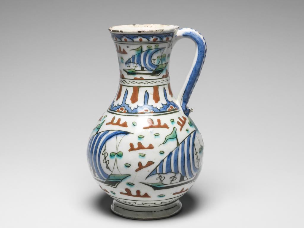 An image of Islamic pottery. Jug. Unknown potter, Turkey, Anatolia, Iznik. Shape: pear shaped body with slightly flared neck sitting on an out-turned foot ring. A strap handle with lower finger rest is attached just below the rim and on the shoulder. Exterior: just below the rim, a frieze of blue lobbed arches is outlined in black and contains a red central dot. On the neck and body, a frieze of lateen-rigged ships with blue and white stripped sails on black and green hulls and rigging float amongst red or green stylised islands, all outlined in black. On these ship friezes, the green pigment has run slightly into the white background. On the shoulder, separating the ship friezes, a band of dashed lines sits above lotus panel arcading in red and blue outlined in black. The handle is painted with blue horizontal and vertical stripes. On the lower body, a frieze of black scrolling lines is enclosed by sets of black concentric lines. Glaze covers the surface except the rim of the foot ring. Interior: surface is coated in glaze but otherwise undecorated. Fritware, probably thrown, coated in a white slip and painted in blue, red, green and black under a transparent white glaze. Height, whole, 22 cm, width, whole, 14 cm, diameter, rim, 8 cm, diameter, base, 8.5 cm, weight, whole, 562 g, circa 1570- circa 1590. Ottoman.