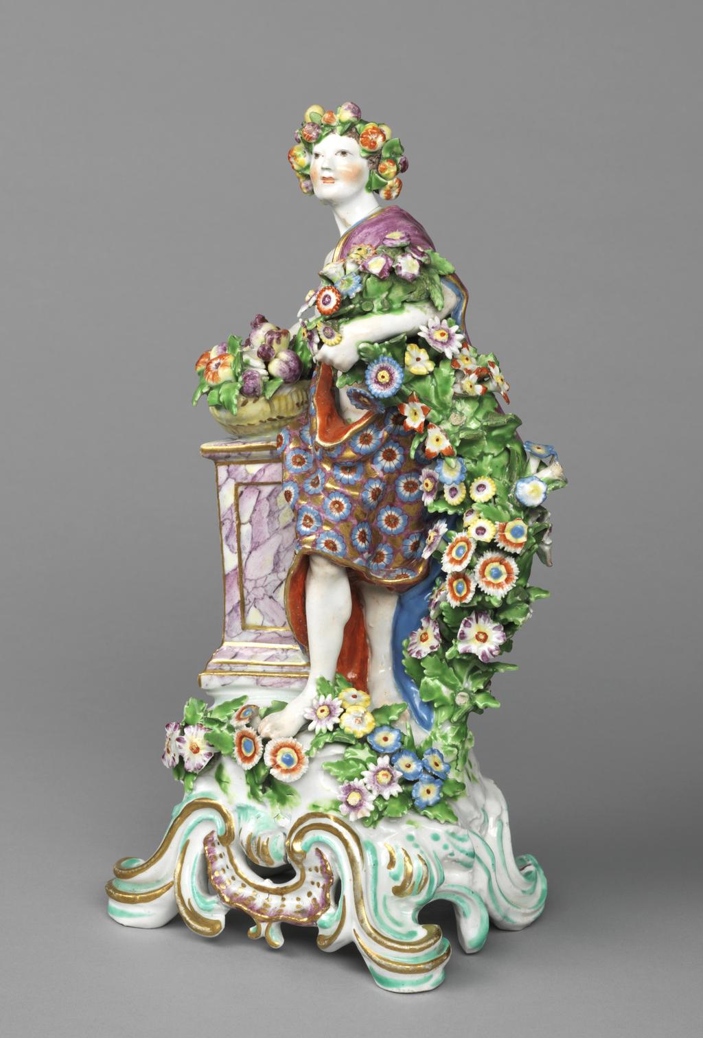 An image of Figure. Youth in Antique Dress, probably representing Autumn. Bow Porcelain Manufactory, Essex, Stratford-le-Bow. The rounded rectangular base has five scroll feet with a pierced rocaille motif between the front two. The front is decorated with applied flowers and leaves. On the viewer's left there is a tall square pedestal on top of which is a basket filled with fruit and foliage. The youth stands beside the pedestal with his right leg in front of his left. His left shoulder is forward and under his left arm he holds up a garland of flowers, which descend to the ground. He leans back slightly on his left arm, which rests on top of the basket. His cheeks are ruddy and he has a wreath of fruit over his black hair. He wears a long drape with a dense pattern of blue and red flowers on a pink and gold ground, and an orange lining, and over it, a pale purple cloak with a blue lining which passes over his left shoulder and down to the ground at the back. The pedestal is marbled in shades of purple, and has gold lines round the panels and edges. The basket is yellow and the fruit and flower garland polychrome. The base is picked out in purple, turquoise and gold. Soft-paste porcelain containing bone ash, press-moulded, with hand-modelled details, painted over lead-glaze in blue, green, yellow, orange, pale purple, and brown enamels, and gilt. The underside has slightly turquoise tinted glaze, and a large central circular ventilation hole. Height, whole, 30.0 cm, width, whole, 16.0 cm, circa 1765-1770. Rococo.
