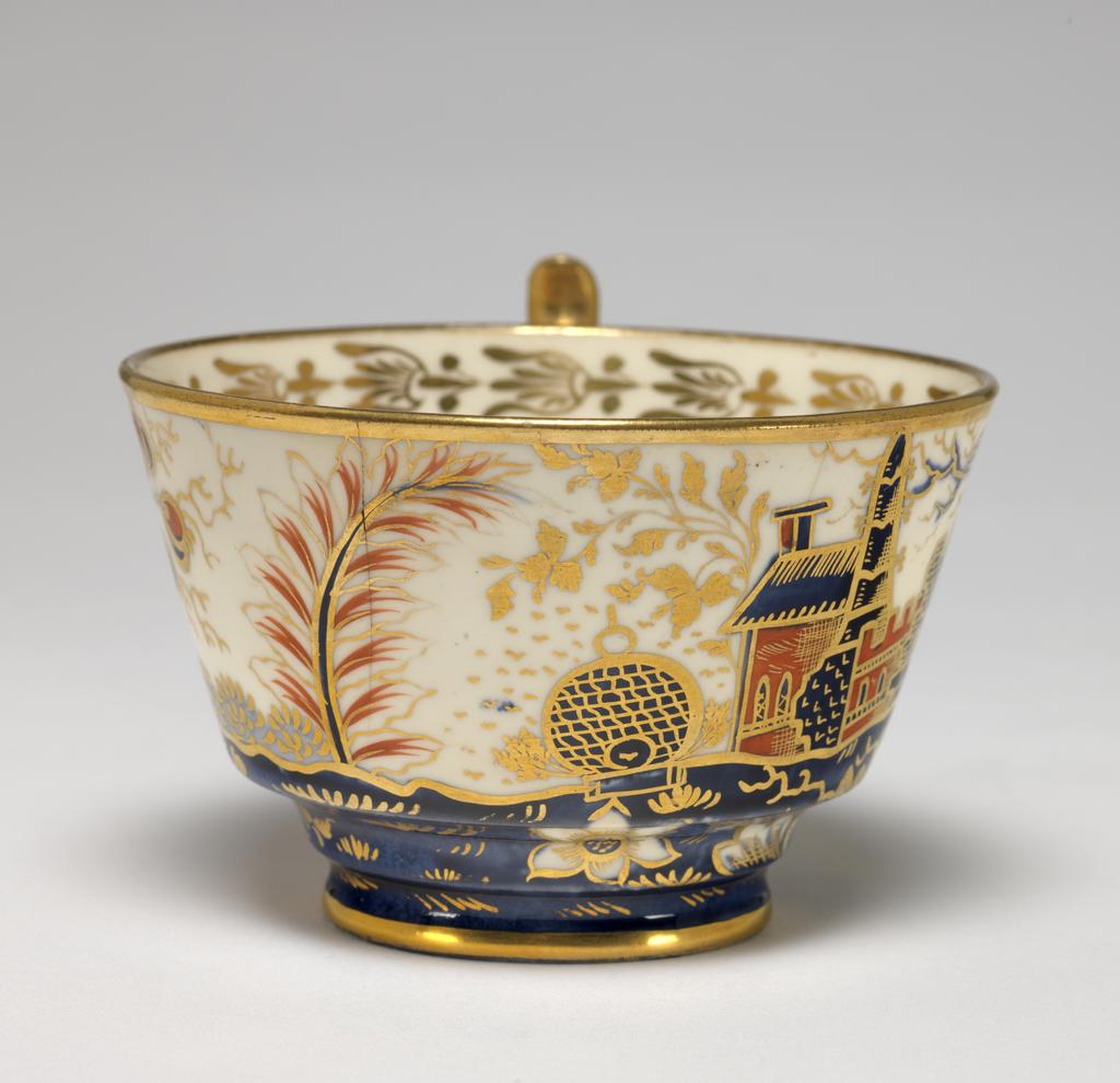 An image of Cup and Saucer/Teacup. New Hall Porcelain Factory, Staffordshire. The cup is of London shape, and the saucer is circular with deep sloping sides, standing on a footring. The exterior of the cup and the interior of the saucer are decorated with a landscape featuring a building with a tower, and a crenellated wing, a ball-shaped beehive, and flanked by stylized trees and bushes, and with a flowering plant in the foreground. The interior of the cup has a gold border of bell flowers with a horizontal band below, gold bands on the edges of rim and foot, and a gilded handle. The saucer has a gold band round the rim. Pattern 570. Bone china? and hybrid hard-paste porcelain, painted underglaze in blue, and overglaze (presumed lead) in red enamel and gilding, height, cup, 6.5 cm, width, cup, 10.5 cm, diameter, cup, 8.9 cm, height, saucer, 3 cm, diameter, saucer, 14.2 cm, circa 1800-1810. Imari Style.