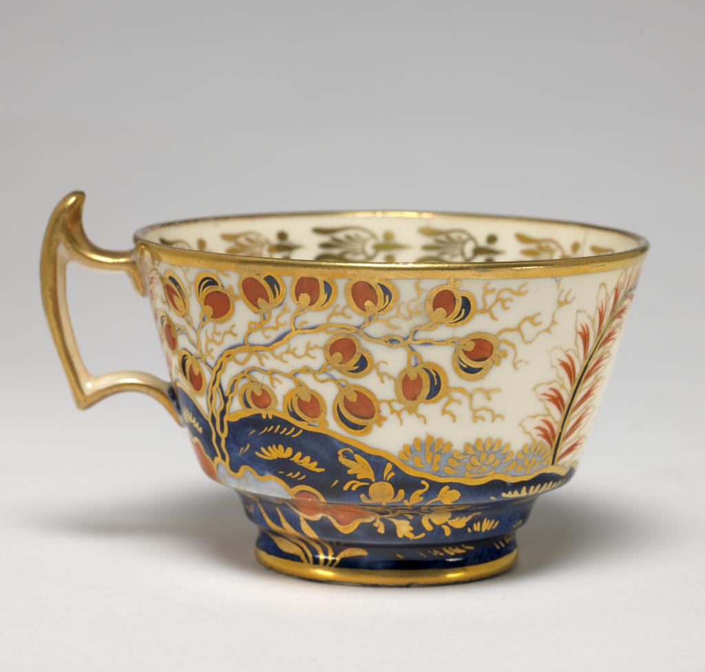 An image of Cup and Saucer/Teacup. New Hall Porcelain Factory, Staffordshire. The cup is of London shape, and the saucer is circular with deep sloping sides, standing on a footring. The exterior of the cup and the interior of the saucer are decorated with a landscape featuring a building with a tower, and a crenellated wing, a ball-shaped beehive, and flanked by stylized trees and bushes, and with a flowering plant in the foreground. The interior of the cup has a gold border of bell flowers with a horizontal band below, gold bands on the edges of rim and foot, and a gilded handle. The saucer has a gold band round the rim. Pattern 570. Bone china? and hybrid hard-paste porcelain, painted underglaze in blue, and overglaze (presumed lead) in red enamel and gilding, height, cup, 6.5 cm, width, cup, 10.5 cm, diameter, cup, 8.9 cm, height, saucer, 3 cm, diameter, saucer, 14.2 cm, circa 1800-1810. Imari Style.