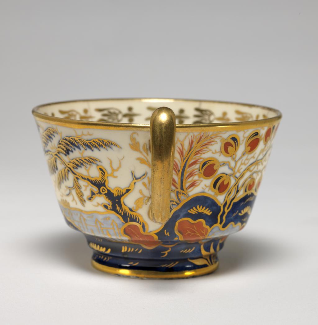 An image of Cup and Saucer/Teacup. New Hall Porcelain Factory, Staffordshire. The cup is of London shape, and the saucer is circular with deep sloping sides, standing on a footring. The exterior of the cup and the interior of the saucer are decorated with a landscape featuring a building with a tower, and a crenellated wing, a ball-shaped beehive, and flanked by stylized trees and bushes, and with a flowering plant in the foreground. The interior of the cup has a gold border of bell flowers with a horizontal band below, gold bands on the edges of rim and foot, and a gilded handle. The saucer has a gold band round the rim. Pattern 570. Bone china? and hybrid hard-paste porcelain, painted underglaze in blue, and overglaze (presumed lead) in red enamel and gilding, height, cup, 6.5 cm, width, cup, 10.5 cm, diameter, cup, 8.9 cm, height, saucer, 3 cm, diameter, saucer, 14.2 cm, circa 1800-1810. Imari Style.