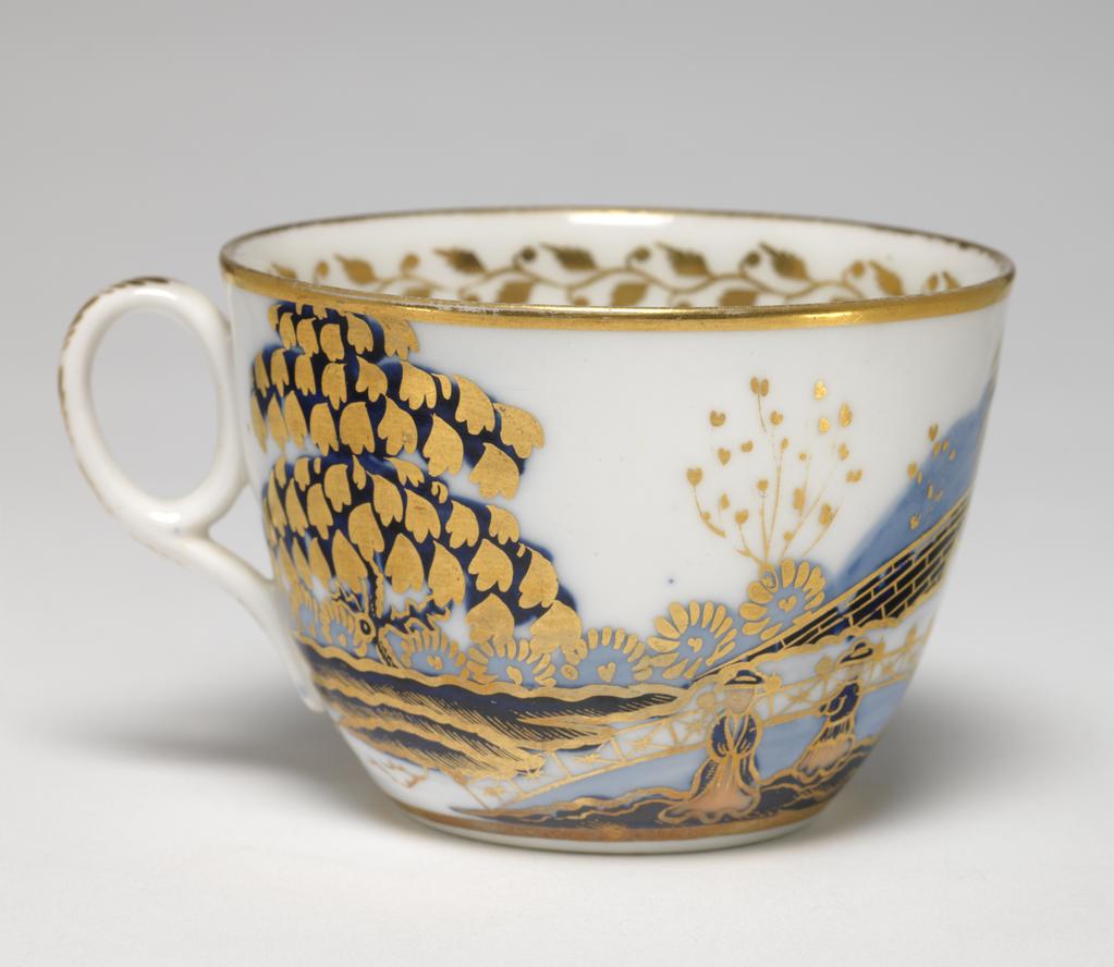 An image of Cup and Saucer/Teacup. New Hall Porcelain Factory, Staffordshire. The cup is of Bute shape with an oval ring handle. The circular saucer is circular with curved sides, standing on a footring. The exterior of the cup is decorated with a landscape with a tree, a man and woman beside a pond, a fence, a large bridge, a house, and a tree. There are gold bands round the base and rim, inside which there is a border of a continuous spray of leaves and buds. At the bottom there is a fruit on a stalk. The back of the handle is decorated with stylized leaves. The saucer is decorated to match with the gold border around it, and a gold band on the rim. Pattern number 1154. Bone china, painted underglaze in blue, overglaze in pale apricot and red enamels, and gilded. Height, cup, 6 cm, width, cup, 10.3 cm, diameter, cup, 8.4 cm, height, saucer, 3 cm, diameter, saucer, 14.2 cm, circa 1815-1820. Imari style.