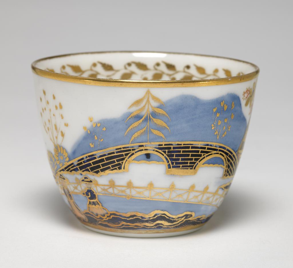 An image of Cup and Saucer/Teacup. New Hall Porcelain Factory, Staffordshire. The cup is of Bute shape with an oval ring handle. The circular saucer is circular with curved sides, standing on a footring. The exterior of the cup is decorated with a landscape with a tree, a man and woman beside a pond, a fence, a large bridge, a house, and a tree. There are gold bands round the base and rim, inside which there is a border of a continuous spray of leaves and buds. At the bottom there is a fruit on a stalk. The back of the handle is decorated with stylized leaves. The saucer is decorated to match with the gold border around it, and a gold band on the rim. Pattern number 1154. Bone china, painted underglaze in blue, overglaze in pale apricot and red enamels, and gilded. Height, cup, 6 cm, width, cup, 10.3 cm, diameter, cup, 8.4 cm, height, saucer, 3 cm, diameter, saucer, 14.2 cm, circa 1815-1820. Imari style.