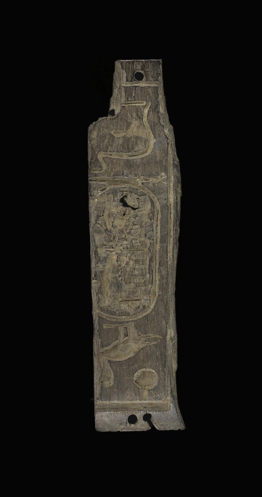 An image of Miscellaneous equipment. Box fragment. Panel with inscription, possibly from a box, with unmodelled back, curved top, with two fixing holes, rectangular bottom with one hold, two holes in right hand edge to take dowels from next panel. Production Place: Egypt. Wood, height 0.125 m, length 0.007 m, width 0.031 m.
