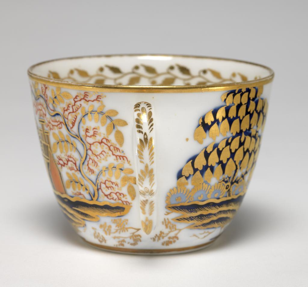 An image of Cup and Saucer/Teacup. New Hall Porcelain Factory, Staffordshire. The cup is of Bute shape with an oval ring handle. The circular saucer is circular with curved sides, standing on a footring. The exterior of the cup is decorated with a landscape with a tree, a man and woman beside a pond, a fence, a large bridge, a house, and a tree. There are gold bands round the base and rim, inside which there is a border of a continuous spray of leaves and buds. At the bottom there is a fruit on a stalk. The back of the handle is decorated with stylized leaves. The saucer is decorated to match with the gold border around it, and a gold band on the rim. Pattern number 1154. Bone china, painted underglaze in blue, overglaze in pale apricot and red enamels, and gilded. Height, cup, 6 cm, width, cup, 10.3 cm, diameter, cup, 8.4 cm, height, saucer, 3 cm, diameter, saucer, 14.2 cm, circa 1815-1820. Imari style.