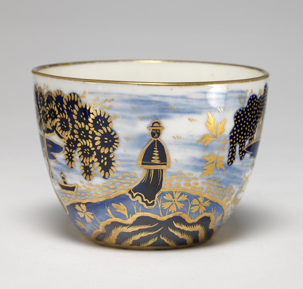 An image of Cup and Saucer/Teacup. New Hall Porcelain Factory, Staffordshire. The cup is of Bute shape with oval ring handle. The saucer is circular with curved sides, standing on a footring. The cup is decorated on the exterior with an Oriental landscape with buildings on the left and right, a man standing fishing on a bridge, a man in a boat, and a larger man in the middle. Inside below the rim there is a continuous horizontal spray of leaves in gold. There is a gold band round the rim, and six stylized leaves down the back of the handle. The interior of the saucer is decorated to match the exterior of the cup and has a gold band round the rim. Pattern 1163. Bone china, painted underglaze in blue, overglaze in apricot-pink enamel, and gilded. Height, cup, 6 cm, width, cup, 10.5 cm, diameter, cup, 8.5 cm, height, saucer, 3.5 cm, diameter, saucer, 14.5 cm, circa 1815-1820. Imari style.