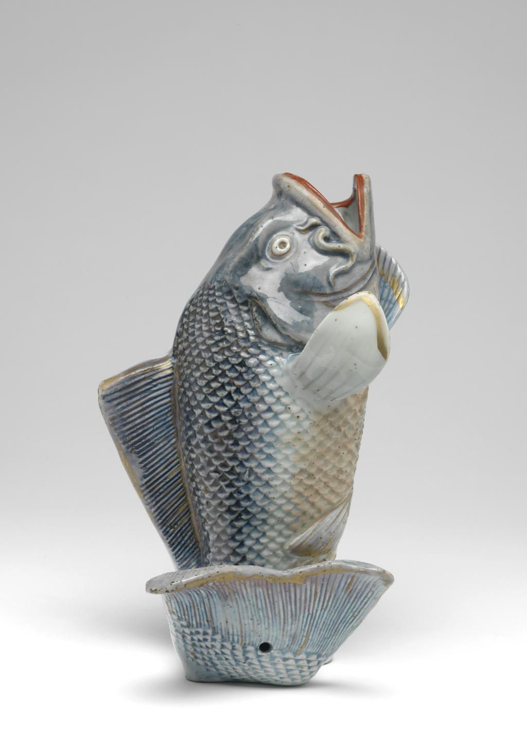 An image of Carp-shaped wall vase. Production Place: Japan, Arita. The carp, hollow-moulded, leaps upwards supported by its curling tail and rests on a simple base with an uneven and gritty underside. The body is finely moulded with scales and gills, and the fins and tail finely incised. The head, upper half of the body, dorsal fin and tail are covered in a wash of underglaze blue, the lower half with pale red enamel and covered with a bluish glaze; details of the tail, fins and head are outlined in gold; the large open mouth is picked out in red and gold and a beady eye in gold with a recessed pupil. The back of the model is flat and undecorated except where the underglaze blue has flowed. A hole is pierced in the centre of the back for suspension and at the base of the tail. Hard-paste porcelain decorated underglaze in blue and overglaze in red, pale-red, gold and violet enamels, and gold, height, whole, 24.2 cm, diameter, whole, 13.3 cm, width, whole, 15.5 cm, circa 1700-1800.