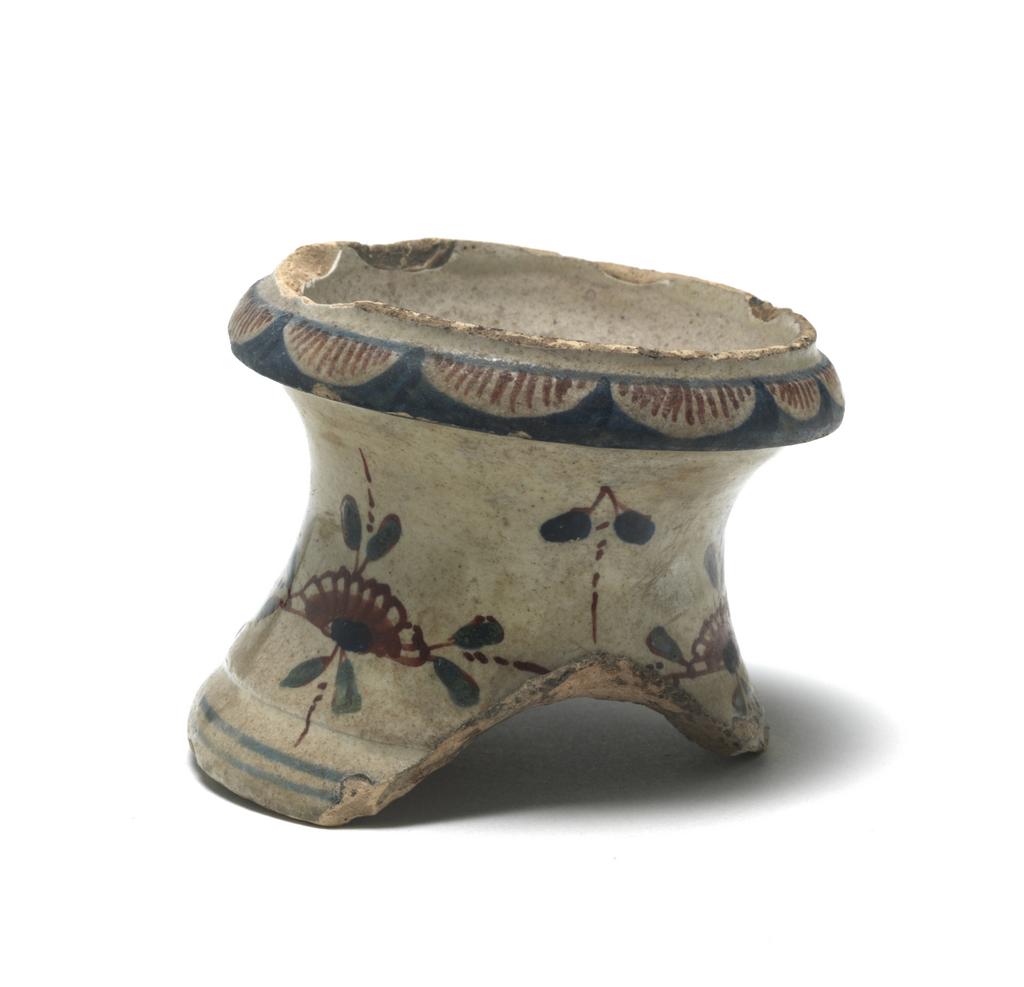 An image of English Delftware. Salt. Part of a salt. Uncertain factory, London, Vauxhall, or Bristol. Of capstan form with an outward curving rim, and a shallow bowl-shaped depression for the salt in the top. About two-thirds of the lower part is missing. The sides are decorated in all three colours with stylized flower and leaf motifs with detached blue flower heads and paired leaves between them. There are two blue bands round the lower edge, and on the rim, a border of semi-circular petals striped in red reserved in a blue ground. The middle of the salt pan is decorated wtih a pair of leaves within two concentric blue circles. Buff earthenware tin-glazed, painted in blue, red, and green, height 5 cm, width, rim 7 cm, circa 1720-1730. English.