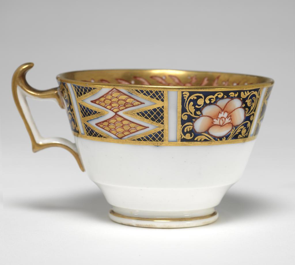 An image of Cup and Saucer/Teacup. New Hall Porcelain Factory, Staffordshire. The cup is of London shape. The circular saucer has deep sloping sides, and stands on a footring. The inside of the cup has a red and black bird on a white area. The rest of the surface is covered with two underglaze blue panels decorated with red and gold flowers, and two white panels decorated with a spray of underglaze blue and green leaves and red flowers, with seven red squares below. Above the panels there are red stylized plants reserved in a gold gound. The exterior is decorated with a border comprising alternating rectangles containing two scale-patterned diamonds reserved in a blue ground, and a red flower reserved in a blue ground. The back of the handle is gilded. The saucer is decorated with the same design as the interior of the cup, with the interior border round the outer edge. Pattern 1368. bone china, painted underglaze in cobalt blue, overglaze in pale green, iron-red, a little black, and gold. Height, cup, 6 cm, width, cup, 10.6 cm, diameter, cup, 8.7 cm, height, saucer, 2.8 cm, diameter, saucer, 14 cm, circa 1813-1818. Imari Style.
