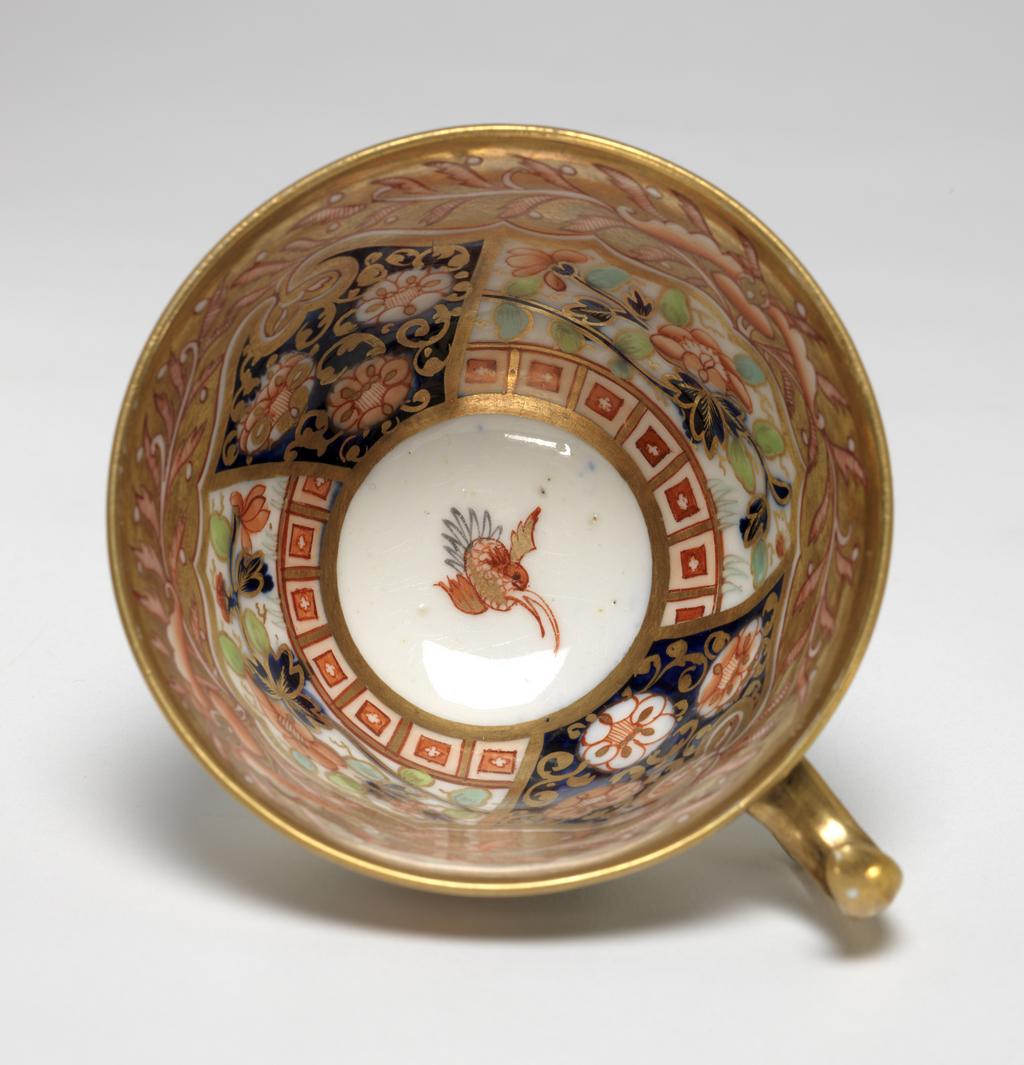 An image of Cup and Saucer/Teacup. New Hall Porcelain Factory, Staffordshire. The cup is of London shape. The circular saucer has deep sloping sides, and stands on a footring. The inside of the cup has a red and black bird on a white area. The rest of the surface is covered with two underglaze blue panels decorated with red and gold flowers, and two white panels decorated with a spray of underglaze blue and green leaves and red flowers, with seven red squares below. Above the panels there are red stylized plants reserved in a gold gound. The exterior is decorated with a border comprising alternating rectangles containing two scale-patterned diamonds reserved in a blue ground, and a red flower reserved in a blue ground. The back of the handle is gilded. The saucer is decorated with the same design as the interior of the cup, with the interior border round the outer edge. Pattern 1368. bone china, painted underglaze in cobalt blue, overglaze in pale green, iron-red, a little black, and gold. Height, cup, 6 cm, width, cup, 10.6 cm, diameter, cup, 8.7 cm, height, saucer, 2.8 cm, diameter, saucer, 14 cm, circa 1813-1818. Imari Style.