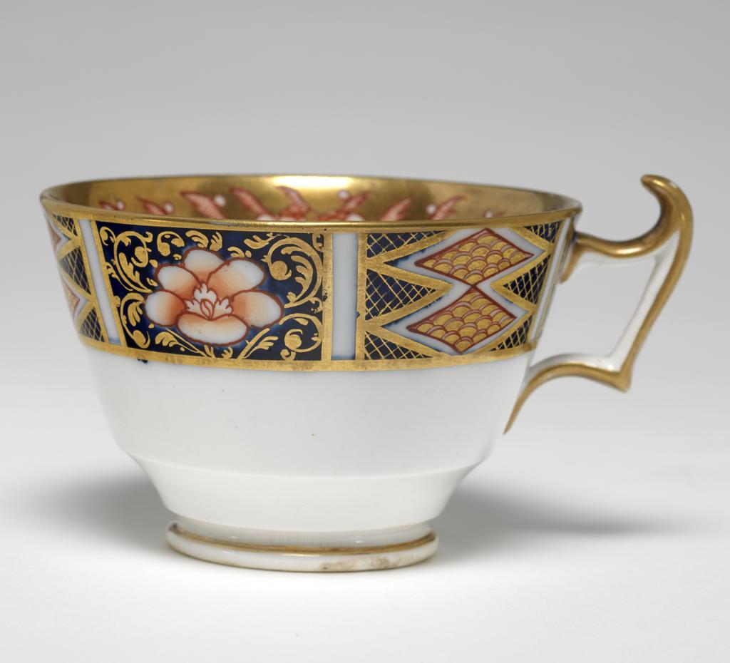 An image of Cup and Saucer/Teacup. New Hall Porcelain Factory, Staffordshire. The cup is of London shape. The circular saucer has deep sloping sides, and stands on a footring. The inside of the cup has a red and black bird on a white area. The rest of the surface is covered with two underglaze blue panels decorated with red and gold flowers, and two white panels decorated with a spray of underglaze blue and green leaves and red flowers, with seven red squares below. Above the panels there are red stylized plants reserved in a gold gound. The exterior is decorated with a border comprising alternating rectangles containing two scale-patterned diamonds reserved in a blue ground, and a red flower reserved in a blue ground. The back of the handle is gilded. The saucer is decorated with the same design as the interior of the cup, with the interior border round the outer edge. Pattern 1368. bone china, painted underglaze in cobalt blue, overglaze in pale green, iron-red, a little black, and gold. Height, cup, 6 cm, width, cup, 10.6 cm, diameter, cup, 8.7 cm, height, saucer, 2.8 cm, diameter, saucer, 14 cm, circa 1813-1818. Imari Style.