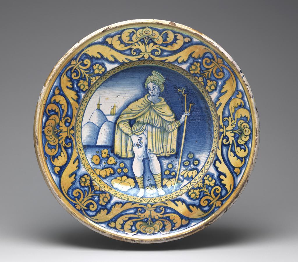 An image of Maiolica. Dish/piatto di pompa. St Roch. Unknown maker, Umbria, Deruta. Shape 61. Circular with slightly sloping rim and wide deep well. standing on a footring pierced by two holes in the correct position for suspension the right way up. In the middle, St Roch leans on a forked staff held in his left hand and points with his right to a plague spot on his right thigh; he has a halo and wears knee-length hose, a short tunic with long sleeves and a cloak. In the background to left, are hills and the towers and spires of two churches, and in the foreground, stylized flowers. The edge of the well has a border of zig-zags and spots. On the rim, there are stylized flowers and scrolling leaves. A band of yellow lustre encircles the outer edge. Dark buff earthenware, the front is tin-glazed; the reverse lead-glazed honey-brown. Painted in bright dark blue and with brassy-yellow lustre, height, whole, 9.0 cm, diameter, whole, 41.8 cm, circa 1500-1550. Renaissance.