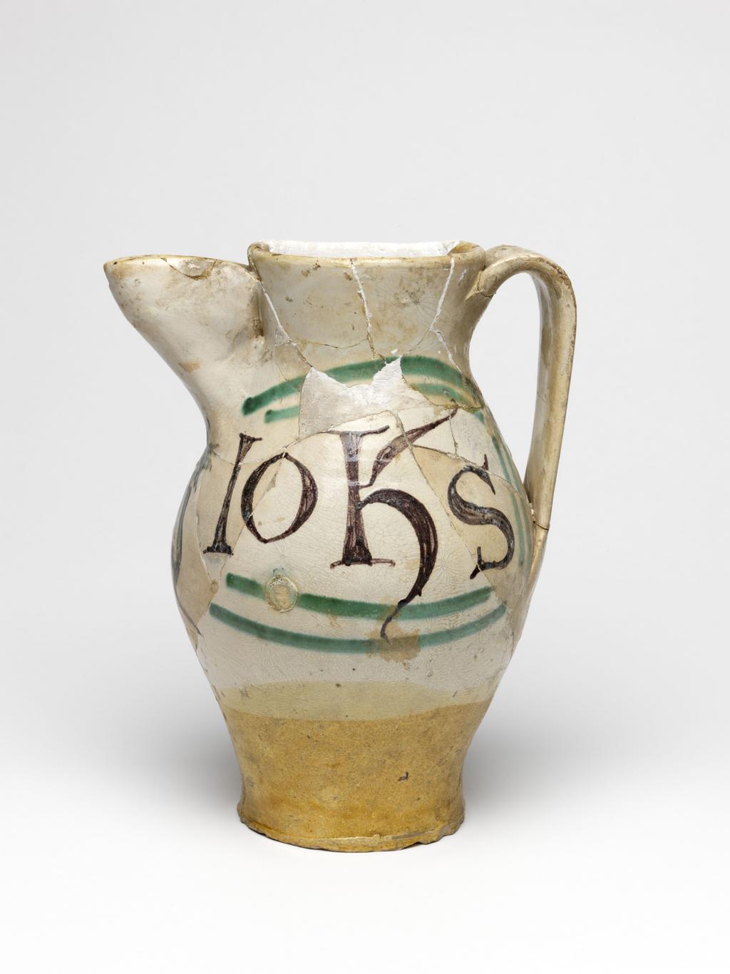 An image of Maiolica arcaica. Jug. Unidentified Orvieto pottery, Italy, Umbria. Shape 7. Ovoid with flat base; cylindrical neck, expanding slightly towards the rim; broad strap handle; spout made from a strip of clay, folded and pressed to the neck. On the front under the spout, a grotesque devil with a striped body, long tail and four horns on its head, faces to the left holding out one of its hands. One side of the jug bears the word `SaS' (Sanctus) in manganese, enclosed by two green lines forming a curved rectangular frame, open on the side nearest the devil. The other side bears the word `IohS' (Johannes), similarly framed. Cream earthenware, the lower part and interior are lead-glazed brownish-yellow, the rest is tin-glazed off-white; the tin-glaze has crazed; painted in manganese and copper-green. Height, whole, 22.7 cm, diameter, base, 8.9 cm, diameter, body, 14.3 cm, width, whole, 17.6 cm, circa 1300-1350. Medieval.