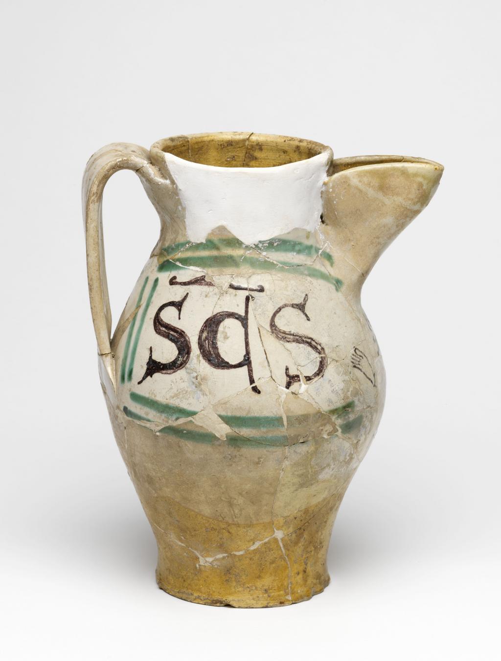 An image of Maiolica arcaica. Jug. Unidentified Orvieto pottery, Italy, Umbria. Shape 7. Ovoid with flat base; cylindrical neck, expanding slightly towards the rim; broad strap handle; spout made from a strip of clay, folded and pressed to the neck. On the front under the spout, a grotesque devil with a striped body, long tail and four horns on its head, faces to the left holding out one of its hands. One side of the jug bears the word `SaS' (Sanctus) in manganese, enclosed by two green lines forming a curved rectangular frame, open on the side nearest the devil. The other side bears the word `IohS' (Johannes), similarly framed. Cream earthenware, the lower part and interior are lead-glazed brownish-yellow, the rest is tin-glazed off-white; the tin-glaze has crazed; painted in manganese and copper-green. Height, whole, 22.7 cm, diameter, base, 8.9 cm, diameter, body, 14.3 cm, width, whole, 17.6 cm, circa 1300-1350. Medieval.