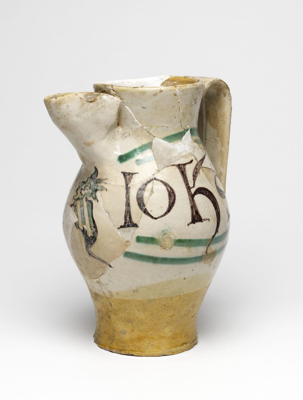 An image of Maiolica arcaica. Jug. Unidentified Orvieto pottery, Italy, Umbria. Shape 7. Ovoid with flat base; cylindrical neck, expanding slightly towards the rim; broad strap handle; spout made from a strip of clay, folded and pressed to the neck. On the front under the spout, a grotesque devil with a striped body, long tail and four horns on its head, faces to the left holding out one of its hands. One side of the jug bears the word `SaS' (Sanctus) in manganese, enclosed by two green lines forming a curved rectangular frame, open on the side nearest the devil. The other side bears the word `IohS' (Johannes), similarly framed. Cream earthenware, the lower part and interior are lead-glazed brownish-yellow, the rest is tin-glazed off-white; the tin-glaze has crazed; painted in manganese and copper-green. Height, whole, 22.7 cm, diameter, base, 8.9 cm, diameter, body, 14.3 cm, width, whole, 17.6 cm, circa 1300-1350. Medieval.