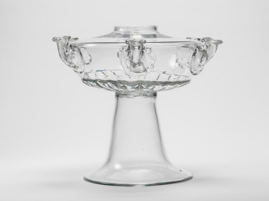 An image of Glass Lamp. Unknown English glassmaker. The depressed globular bowl of the lamp is blown-moulded with gadrooning round the lower part, six applied nozzles for wicks at regular intervals on the sides, and a circular hole in the centre. It is joined to a tall, plain inverted trumpet-shaped foot. Clear lead glass, lamp bowl blown-moulded, nozzles applied decoration, height, whole, 22.0 cm, diameter, foot, 15.7 cm, circa 1700-1725. Baroque. Queen Anne, George I.