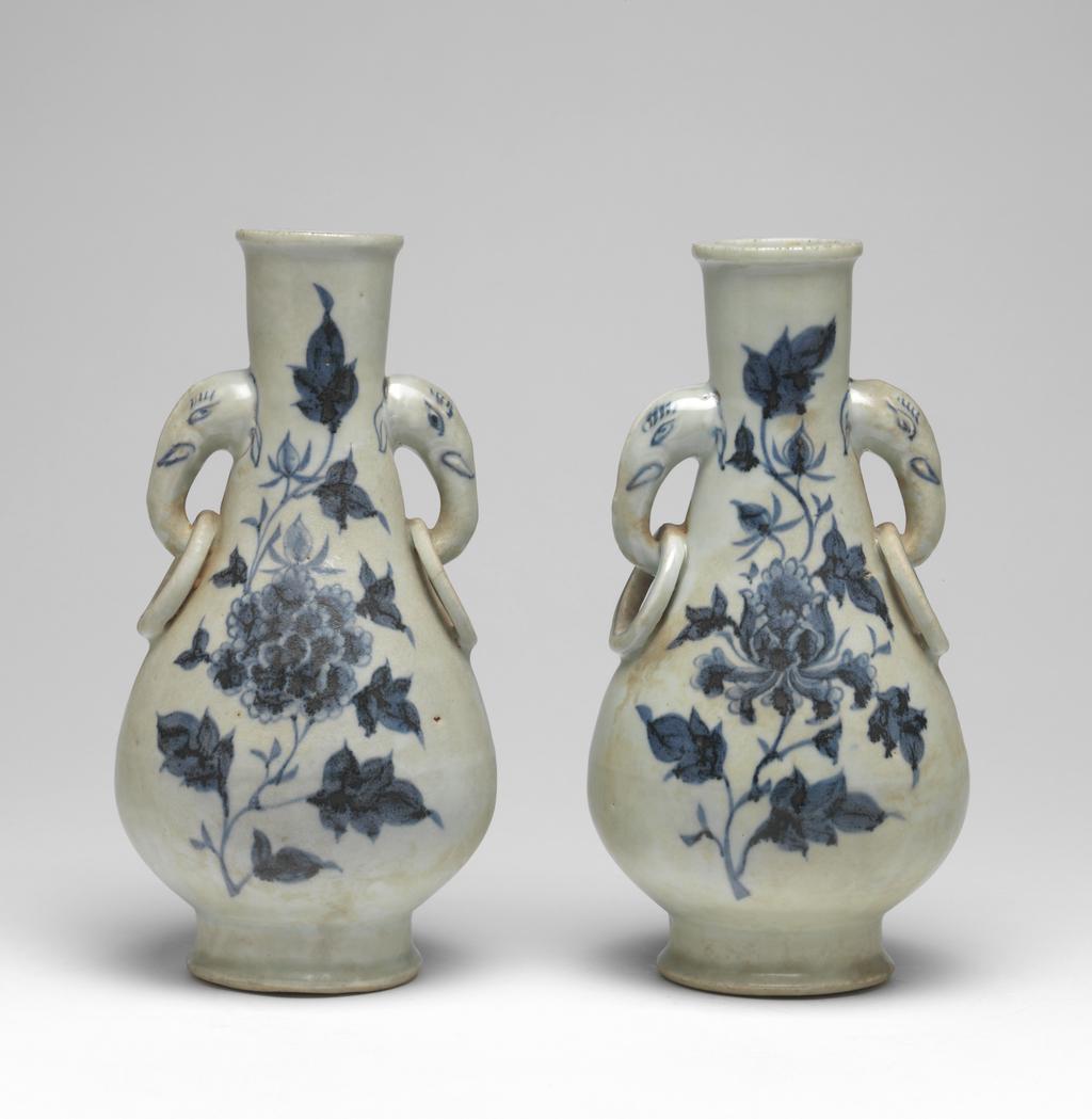 An image of Vase with elephant handles. Unknown maker, China, Folk kilns. The pear-shaped vase has a slender neck, thick, rounded lip, and two elephant head and fixed-ring handles. The body tapers to a deep, flared foot with deep flat, recessed base. The elephant handles are finely painted with eyes, eyelashes, tusks and ears, and the fixed rings with single circle ornament. On each side of the body are elegant peony sprays in blue. The interior is unglazed, as is the base which is covered with a red, sandy deposit. Hard-paste porcelain, thrown, painted in underglaze blue, with base and interior unglazed. Height, whole, 24.6 cm, diameter, whole, 12.7 cm, circa 1300-1368. Yuan Dynasty (1206-1368). Collection: H.S. Reitlinger. Of a pair of vases, with C.689B-1991.