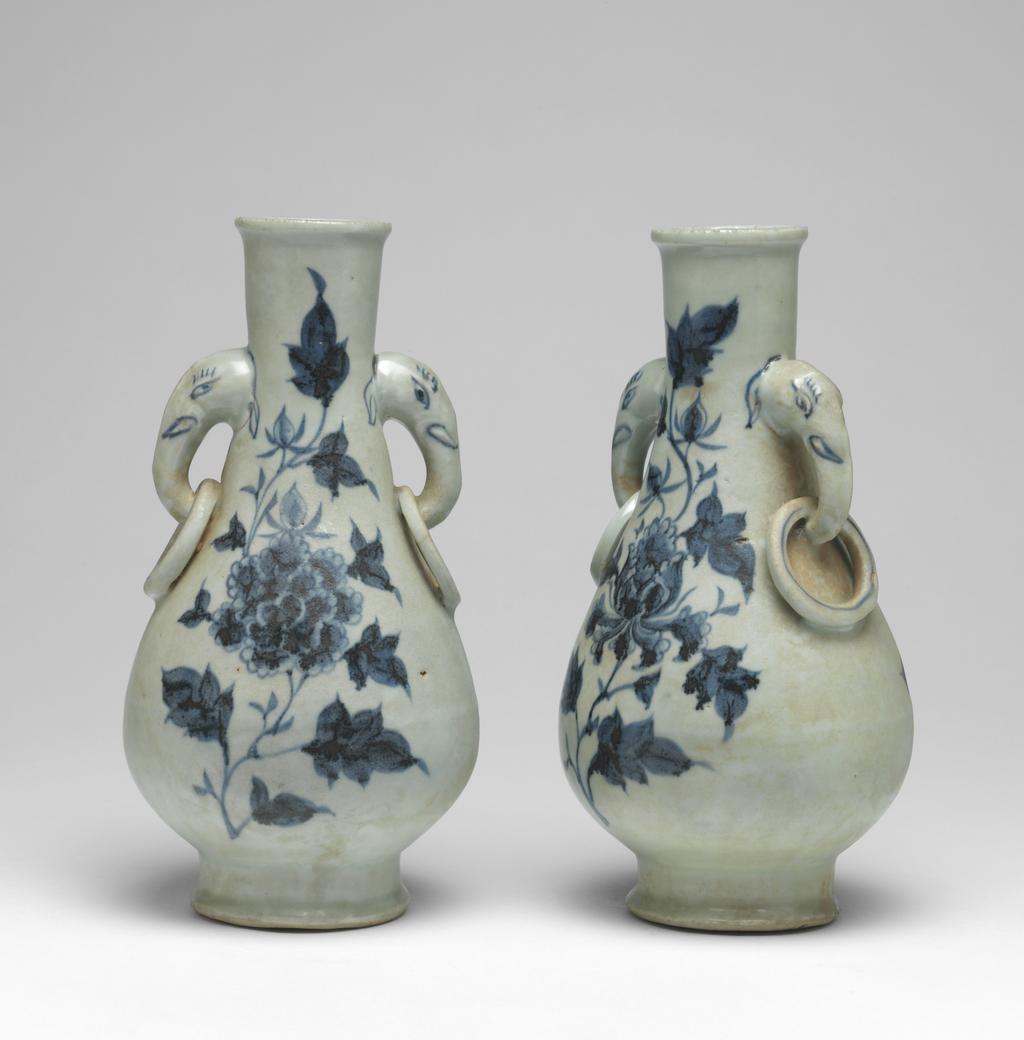 An image of Vase with elephant handles. Unknown maker, China, Folk kilns. The pear-shaped vase has a slender neck, thick, rounded lip, and two elephant head and fixed-ring handles. The body tapers to a deep, flared foot with deep flat, recessed base. The elephant handles are finely painted with eyes, eyelashes, tusks and ears, and the fixed rings with single circle ornament. On each side of the body are elegant peony sprays in blue. The interior is unglazed, as is the base which is covered with a red, sandy deposit. Hard-paste porcelain, thrown, painted in underglaze blue, with base and interior unglazed. Height, whole, 24.6 cm, diameter, whole, 12.6 cm, circa 1300-1368. Yuan Dynasty (1206-1368). Collection: H.S. Reitlinger. Of a pair of vases, with C.689A-1991.