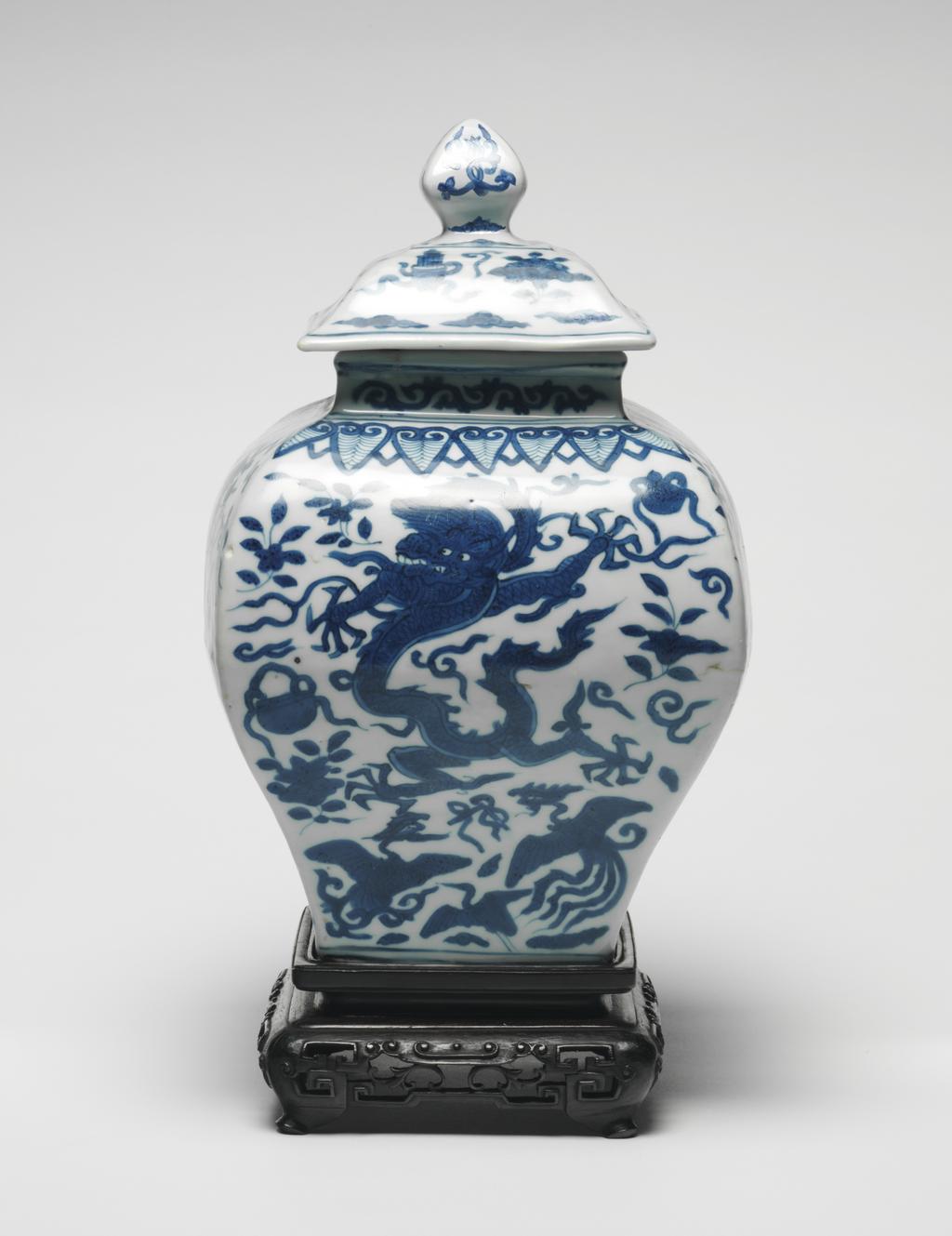 An image of Square section baluster jar and cover with wooden stand. The jar is painted on all sides with five-claw dragons, phoenixes and the eight precious emblems below a border of ruyi head lappets around the shoulders and scrolls on the neck. The cover is painted with the eight precious emblems and cloud scrolls. Hard-paste porcelain, thrown, decorated in underglaze-blue, height, jar, 26.6 cm, height, whole, 32.0 cm, diameter, whole, 17.6 cm, circa 1573-1619. Ming Dynasty (1368-1644), Wanli period (1573-1619). Chinese. As a pair with C.716.2 & A & B-1991.