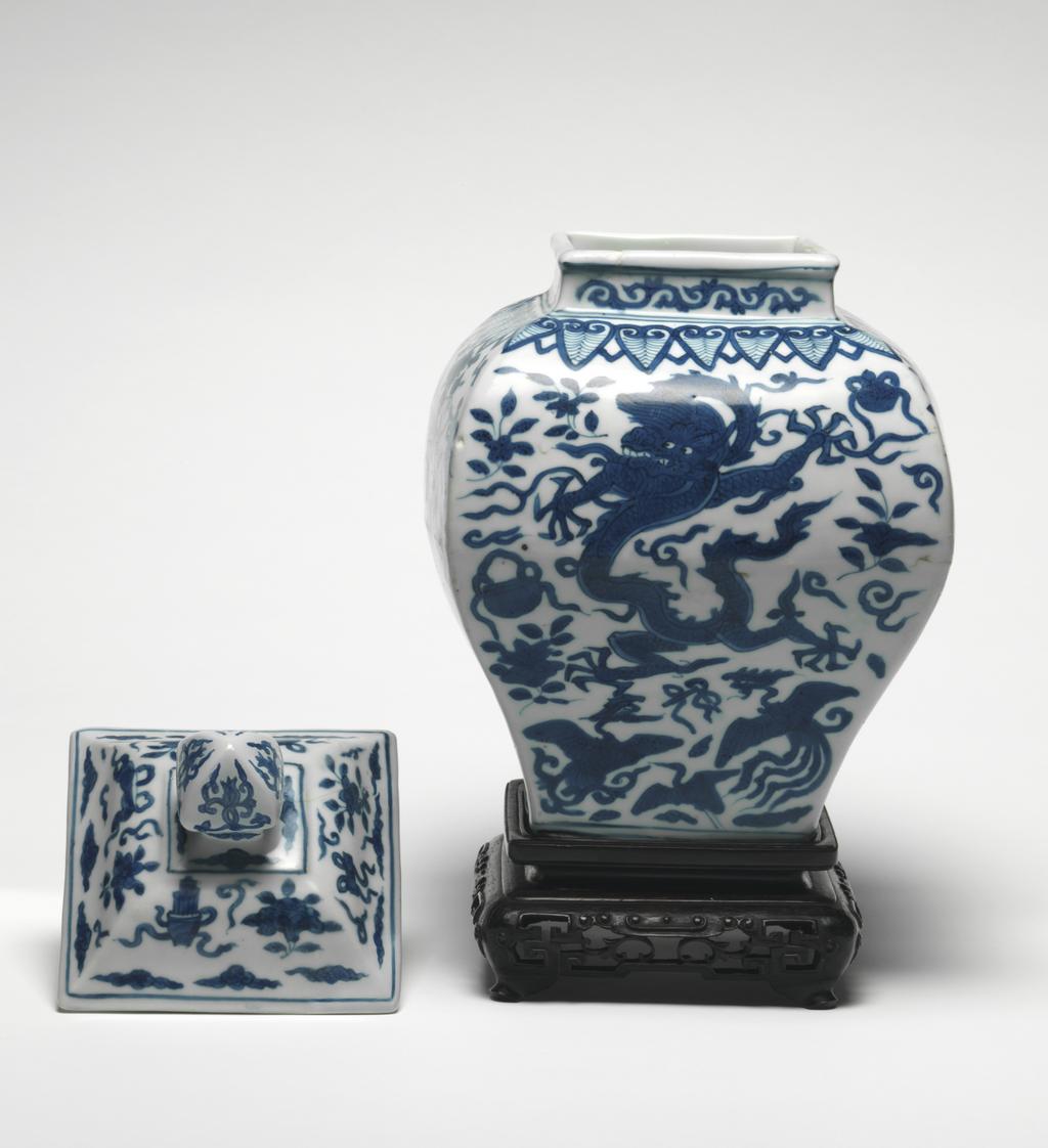 An image of Square section baluster jar and cover with wooden stand. The jar is painted on all sides with five-claw dragons, phoenixes and the eight precious emblems below a border of ruyi head lappets around the shoulders and scrolls on the neck. The cover is painted with the eight precious emblems and cloud scrolls. Hard-paste porcelain, thrown, decorated in underglaze-blue, height, jar, 26.6 cm, height, whole, 32.0 cm, diameter, whole, 17.6 cm, circa 1573-1619. Ming Dynasty (1368-1644), Wanli period (1573-1619). Chinese. As a pair with C.716.2 & A & B-1991.