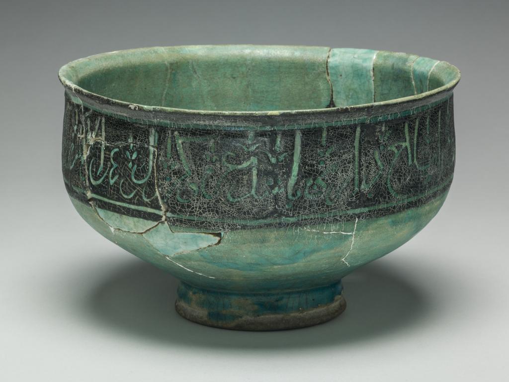 An image of Bowl. Islamic pottery. Silhouette ware. Unknown potter, Iran. Shape: irregular carinated bowl with conical lower body and cylindrical neck with an everted rim. Sits on a foot ring. Interior: on the body four black palmettes toped with finials are spaced at regular intervals, with an additional, smaller version at the centre of the base. Glaze pools in the base. Exterior: On the neck a wide band of black is incised with script with fleurs de lys at regular intervals. Glaze runs terminates on the foot ring, where coverage is uneven. Fritware, wheel thrown, partially coated in a black slip, carved under a transparent, crazed turquoise glaze. Height, whole, 12 cm, width, whole, 19.7 cm, diameter, base, 8.2 cm, weight 587 g, circa 1100-circa 1199. Seljuk.