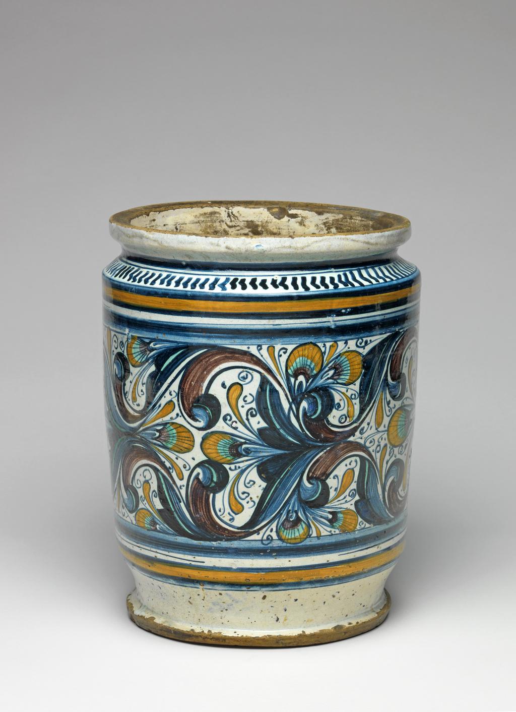 An image of Maiolica. Pharmacy jar/albarello. Unidentified Pesaro potter, The Marches. Broad albarello with straight sides. On the front, in a contour panel with three lobes on each side, are profile busts of a man and woman wearing contemporary costume, facing inwards towards a vase of pinks, with groups of three dots in the background. The rest of the main field is decorated with gothic foliage and peacock's feather eye motifs. Above and below there is a wide blue band and a dark yellow band between two blue; on the shoulder, a row of blue chevrons with a horizontal blue band above. `4S758' is incised into the base. Buff earthenware, tin-glazed on the exterior and more thinly on the interior; base and outer edge of base unglazed. Painted in dark blue, green, dark yellow, and manganese-purple. Height, whole, 26.9 cm, diameter, rim, 20.0 cm, diameter, base, 17.3 cm, diameter, whole, 21.2 cm, circa 1470-1480. Renaissance.