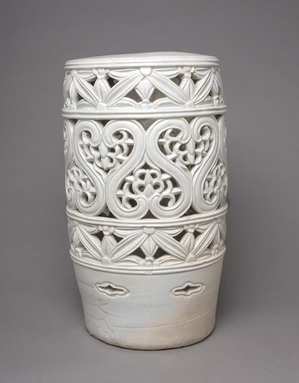 An image of Stand with floral designs. Unknown pottery, Korea. This large stand is cylindrical and has a hole in the top. The sides are divided into three horizontal zones and decorated with two bands of pierced floral designs reminiscent of Korean latticework, around a wider central pierced band with unusual S-shaped motifs enclosing leaves; above the base are small eye-shaped (ansang) openings. The glaze has a pale bluish tone. The stand has no base and the footring shows remains of coarse sand from the firing support. Porcelain, thrown, carved, pierced and glazed, height, whole, 48.7 cm, diameter, top, 26.3 cm, diameter, foot, 23.5 cm, circa 1800-1900. Choson Dynasty (1392-1910). Production Note: This piece was probably used as a stand for a flowerpot.