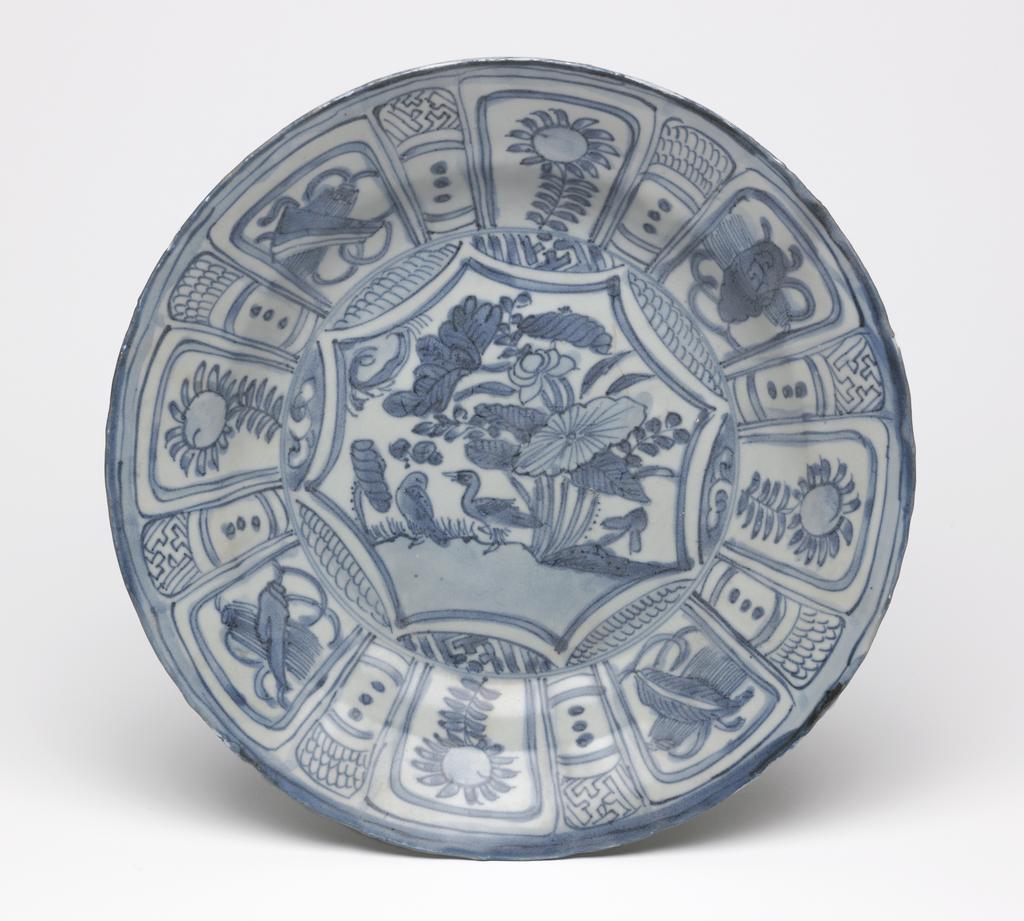 An image of Dish/Kraak ware. Unknown production, China. This dish has a slightly rounded, shallow cavetto. The wall above the cavetto slants outward to form a wide, flattened mouth with a foliated rim. In the centre of the dish decorated with ducks next to a lotus pond and the panels of the rim are subdivided into four ornamental pairs that face each other across the dish. Cobalt blue was applied throughout to outline the motifs, which were then coloured with shades of blue typical of the Wanli style, but the glaze is degraded. A spare linear design of simplified panels and bands with tassels is sketched on the exterior. Hard-paste porcelain, painted underglaze in cobalt blue, height, whole, 5 cm, diameter, whole, 27.5 cm. Wanli Period (1573-1620).