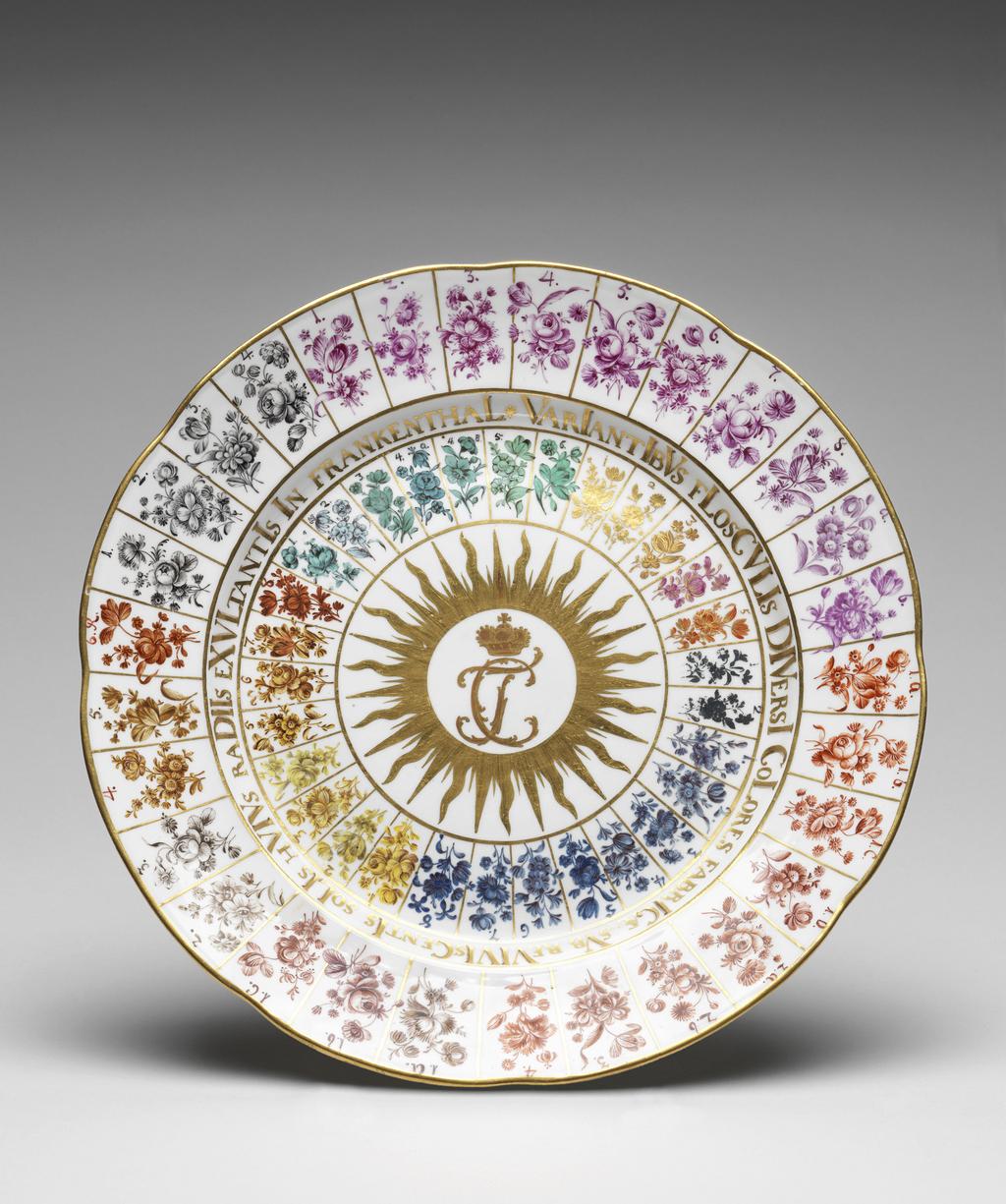 An image of Trial plate. Frankenthal Porcelain Factory, Germany, The Palatinate. Circular with eight slight arcs on the edge, sloping concave rim, shallow curved sides, and flat centre, standing on a footring. Decorated in the centre with a gold crown of TC monogram (Carl Theodore) surrounded by a sunburst with wavy rays. Around this are thirty radiating compartmens outlined in gold, each painted with a spray of flowers. On the sides of the well is an inscription in Latin VARIANTIBVS FLOSCVLIS DIVERSIL COLORES FABRICA SVB REVIVI GENTIS SOLIS HVIVS RADIIS EXVLTANTIS IN FRANKENTAAL. Hard-paste porcelain, moulded, painted in enamels, and gilded, height 3.2 cm, diameter 24.7 cm, circa 1775. Rococo. Notes: Carl Theodore was the Elector Palatine.