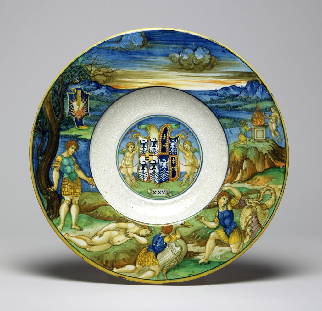 An image of Maiolica. Dish from the Isabella d'Este Service. Nicola di Gabriele Sbraghe da Urbino (Italian, d.1537/38). Shape 56. circular with broad, almost flat rim, and deep well; the underside of the rim moulded with three bands of reeding. In the well, within narrow blue and yellow concentric circles, two putti support a shield charged with the arms of Gonzaga impaling Este: argent, a cross patty gules cantoned by four eagles displayed sable (Gonzaga modern granted 1433), an escutcheon overall, quarterly, 1, 4, gules a lion rampant argent (Bohemia), 2, 3, barry of six or and sable (Gonzaga ancient) impaling Este, quarterly, 1, 4, azure three fleurs-de-lis or a bordure indented gules, 2, 3, azure an eagle displayed argent (gules shown orange throughout). Below on a scroll is the impresa `XXVII'. The sides are decorated in bianco sopra bianco with palmettes and scrolls, and a blue line encircles the shoulder. The rim is decorated with Peleus and Thetis. Peleus stands beneath a tree, gazing at Thetis, who sleeps nude on the ground in front of him. Further to the right she is transformed into a swan-like bird as he embraces her, and then into a dragon from which he turns away in horror. Higher up on the right, a goddess appears to Peleus who kneels before an altar. In the distance there is a mountainous landscape and sky. Suspended from the tree there is a shield of testa di cavallo form charged with the impresa of gold rods in a crucible surrounded by flames in a blue ground. Earthenware, tin-glazed overall; the reverse pale beige and speckled. Painted in blue, green, yellow, orange, stone, brown, manganese-purple, black, and white high-temperature (metallic oxide) colours, height, whole, 3.9 cm, diameter, rim, 30.2 cm, circa 1524-1525. Renaissance. Notes: The figures were derived from a woodcut illustration in Giovanni dei Bonsignori's Ovidio methamorphoseos vulgare, Venice (Giovanni Rosso da Vercelli for Lucantonio Giunta), 1497 or a later edition.