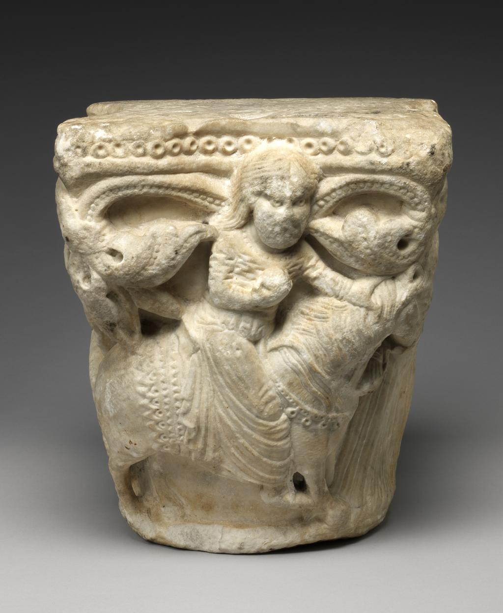 An image of Sculpture. Capital of a column, the sides carved with a man seated on a throne playing the harp, probably the psalmist, David, with an eagle above and to the right of him, a woman riding a horse, possibly the Virgin Mary, and another woman leading the horse with her right hand and holding an eagle on her left. Round the upper edge there is a row of drilled beading with below, foliated scrolls flanking the figures. White marble, carved and drilled, height, whole, 29.8 cm.  Production Place: France, Avignon. Probably from the Romanesque cloister of Notre-Dame-des-Doms, Avignon. Circa 1150-1200. Romanesque.