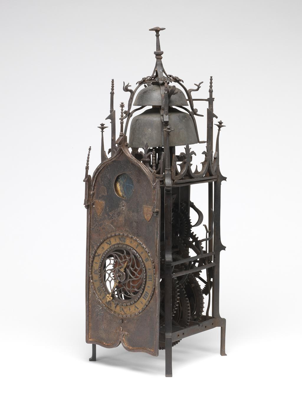 An image of Metalwork. Gothic Iron Clock. Unknown clockmaker. Probably German, but the clock bears some resemblance to the Swiss Liechti clocks of the sixteenth century. Iron clock with weight driven movement of fifteen hour duration and striking hours and quarters. Dial. Ogee arched-top dial plate with pierced tracery centre and chapter ring between two bands of simple Greek fret. The white painted chapter ring (now discoloured) with Roman numerals I-XII, and simple half hour markings in black. Pierced and shaped gilt single iron hand which may be of later date. In the arch, a circular aperture with a rotating disc behind to show the phases of the moon. At the base of the arch are two small raised shields with indistinct quarterings. Movement. Weight driven movement, of fifteen hour duration, and striking hours and quarters. The whole clock made of iron. The going train controlled by a verge escapement and foliot balance with moveable square weights for adjusting the timekeeping. The quarter striking train in the middle of the frame, with a vertical hammer arbor and strikes on the larger bell. Release of the striking is controlled by a nag's head lifting piece, so no warning action occurs. Spare holes in the bottom bars of the frame have been made later to increase the duration between winding by suspending the weights on pulleys and looping up the ends of the cords. The frame has typical Gothic decoration in the iron work and, as is normal with these clocks, there is no provision for enclosing the movement. There has been qjite a lot of repainting of the clock. Iron, height, whole, 53.3 cm, width, whole, 16.8 cm, depth, whole, 20.4 cm, circa 1500-1550.