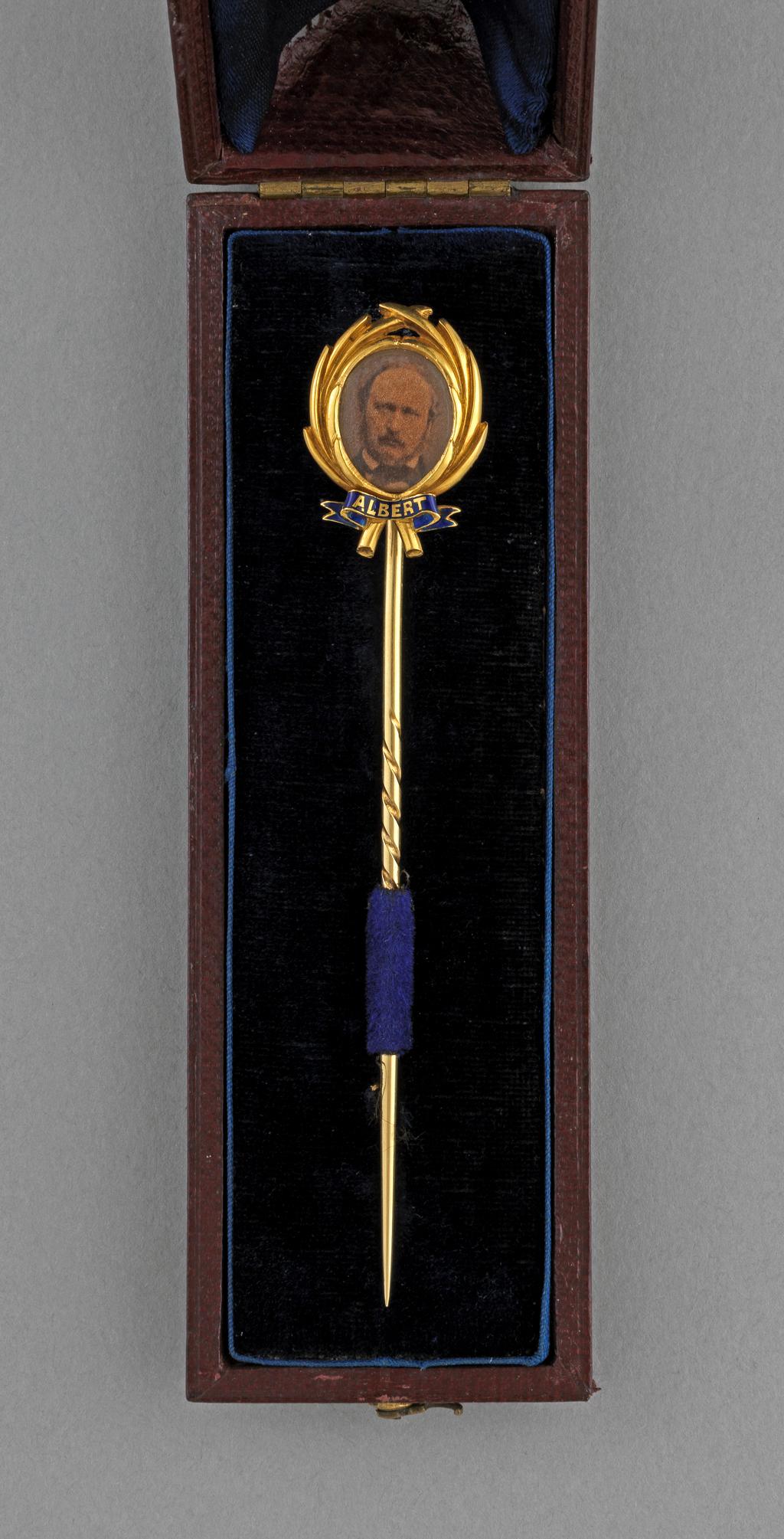 An image of Stick-pin. Gold, the head in the shape of a wreath framing an oval photograph of Prince Albert under glass, the name 'ALBERT' reserved in gold in a blue enamel ribbon beneath. The back inscribed 'To Lord George Lennox/In remembrance/of the/beloved Prince./Decr.14.1861./from V.R.' In original maroon leather case (A) lined with royal blue velvet and silk. Height (whole) 9.5 cm, width (whole) 1.8 cm, after 1861.