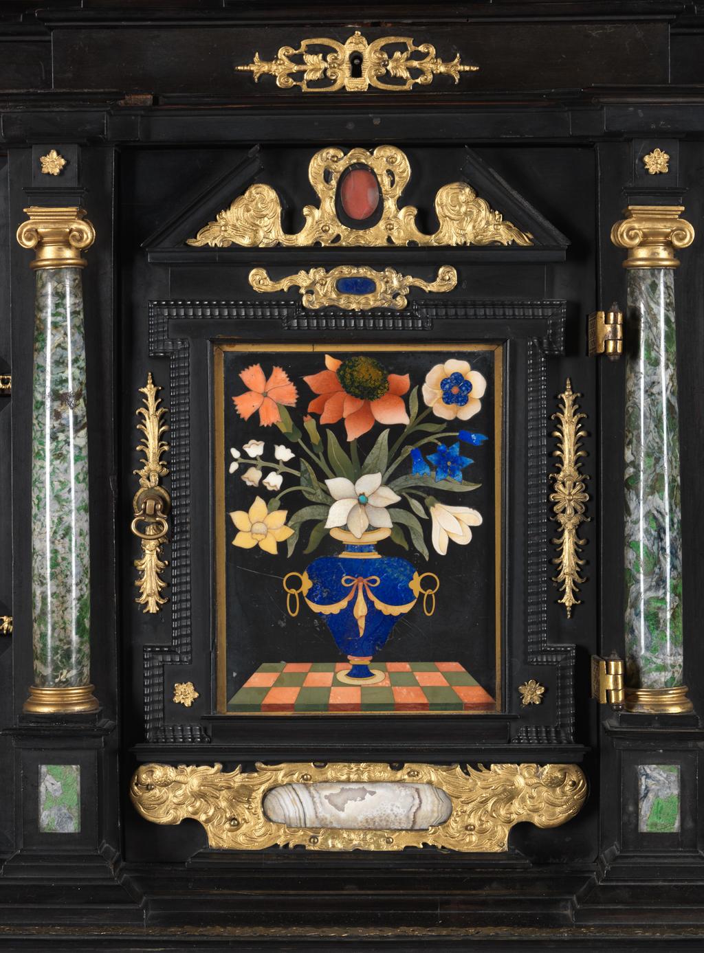 An image of Augsburg 17th century silver-gilt mounted ebony Table Cabinet with Florentine pietre dure plaques from the Grand Ducal workshops. Boscher, Elias (1600-1674). Of architectural form, the stepped superstructure fitted with three frieze drawers above a central cupboard door, architectural pediment and inset with a panel depicting a chaffinch on a fruiting bough, flanked by columns of Smaragdite Gabbro with Corinthian capitals flanked by two deep drawers, inset with panels depicting parrots within fruiting boughs, with three further fitted drawers below, the large central cupboard door inset with a panel of a lapis lazuli flower filled urn issuing stems including a carnation, anemone, daffodil and lilies and enclosing an architectural interior, flanked by further Smaragdite Gabbro Ionic columns and three drawers to each side inset with further symmetrical floral panels including tulips, roses, carnations, lilies and bluebells, on shaped bracket feet, each side inset with large central panels of parrots on fruiting boughs, with four further floral panels above and below, the reverse veneered in kingwood, amaranth and stained walnut in geometric stellar designs. The cabinet containing a complex series of fourty-four secret drawers and compartments to the interior. The cabinet is signed in pencil on the underside 'Elias Boscher' and 'gemacht'. The central cabochon mount stamped with the Augsburg town mark and the silversmiths mark of Johann Spitzmacher (active 1655-1678). The front bears the ebony stamp "EBEN" with the pinecone of Augsburg. Height 85 cm, width 84cm, depth 40cm, circa 1660.