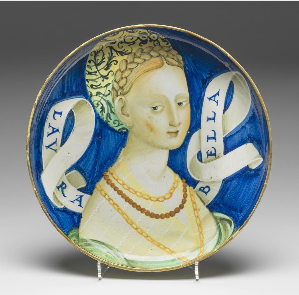 An image of Maiolica. Shallow bowl on low foot. Unidentified potter. This type of dish is traditionally attributed to Castel Durante, but could have been made in Urbino. A bust of a young woman, three-quarters to right, her gaze cast downwards to her right. Her blonde hair is dressed in a plait over the crown of her head. She wears a green and yellow turban embroidered with black scrolling foliage; two gold chains and a rope of beads; and a green dress with a yellow chemise showing on the shoulders. A scroll inscribed `LAVRA/BELLA' in blue projects on either side of her head. The background is blue. On the back are four concentric yellow bands: on the edge of the foot, just beyond the junction of the foot and bowl, and two round the rim. Pale buff earthenware, tin-glazed overall. Painted in blue, green, yellow, orange, cream, and black. Height, whole, 4.7 cm, diameter, whole, 22.7 cm, circa 1525-1535. Renaissance.