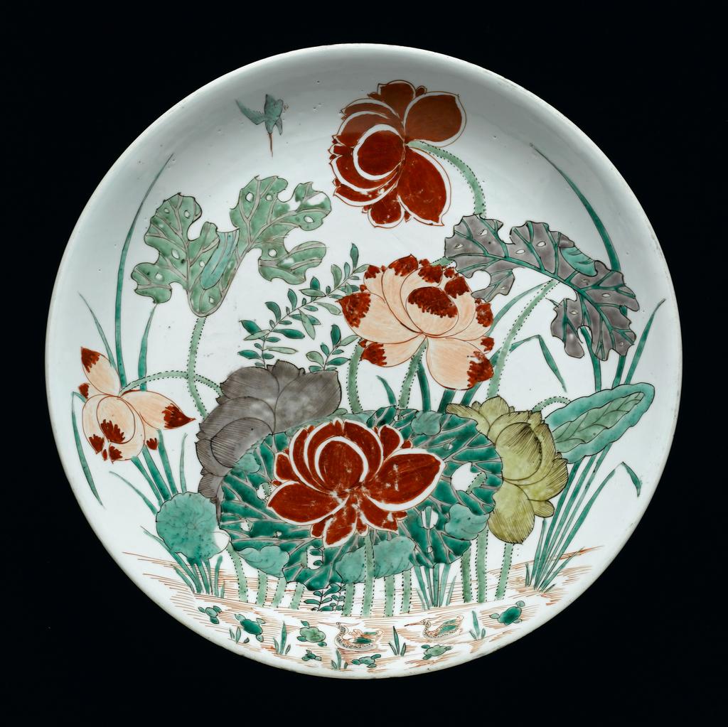 An image of Description: Saucer dish. Hard-paste porcelain painted in an enamel palette of two iron-reds, two greens, turquoise, pale yellow and aubergine outlined in sepia with a kingfisher in flight by a lotus pond with two mandarin ducks swimming.Saucer dish. Hard-paste porcelain painted in an enamel palette of two iron-reds, two greens, turquoise, pale yellow and aubergine outlined in sepia. The well rounded sides have an out-curved rim resting on a low tapered foot. The interior is boldy decorated with an overall design of a kingfisher diving down towards a large clump of lotus with six open blooms, flanked by frilly-edged leaves and tall water grasses, rising from a pond indicated by iron-red dashes, with two mandarin ducks swimming in the foreground. The base has two concentric circles in underglaze blue enclosing the mark. Production Place: China (country) Dimensions: diameter: (whole): 39 cm Period : Qing Dynasty (1644-1912), Kangxi Period (1662-1722) Date: circa 1662 to 1722   