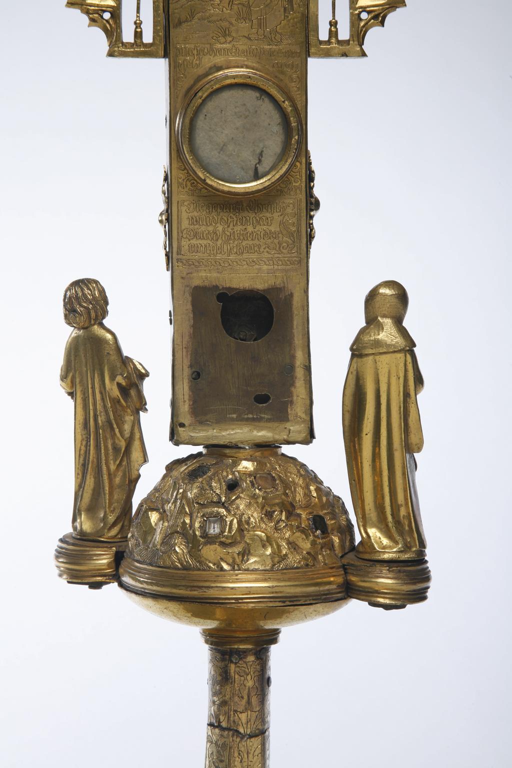 An image of Sculpture. Crucifix/altar cross. Sexfoil engraved with the Resurrection; the angel with the two Maries at the tomb; Noli me tangere; the walk to Emmaeus; the Ascension; and Pentecost. The cross, which contains a reliquary, is set with imitation stones, and enclosed in canopy work among which are the figures of the twelve apostles, and the central figure of the donor, or the saint to which the church was dedicated. There is an angel at either end of the cross-bar, both holding a chalice. There are four circular medallions of enamel on the front of the cross. Top: St. Luke with the bull; right St. Mark with the lion; left: St. John with the eagle; bottom: Adam and Eve expelled from the garden of Eden. (?St. Matthew at the top and missing, as the plaque of the Expulsion should be on the reverse of the cross). Beneath the Expulsion are the words: 'Johanesz hieber diener/meinn. Lasz dir meinn/mütter befollen seinn.' (John, I herewith commend my mother to you.') which refers to the figures of the Virgin Mary and St. John who are standing at the foot of the cross. [The Virgin on the left and St. John on the right]. The three enamel medallions on the back of the cross are; top: Creation of Eve from Adam's side; left: Eve tempting Adam with the apple; right: Adam and Eve hiding from the Lord God in the garden. The fourth medallion is missing, and it may be suggested that the Expulsion should belong here. Engraved at the top of the back of the cross is God the Father and Creator, crowned with a Jewish high-priestly mitre (or the imperial crown?), and holding an orb and a sceptre terminating in a fleur-de-lis. Below, on the cross-bar, is the garden of Eden with many animals (which perhaps represent unfallen nature). Below this again is the scene from Numbers XXI. Here a man lies bitten by a snake, in front of the tents of the camp of the Israelites, while Moses, with two horns upon his head stands by. There is a reliquary compartment behind the figure of Christ, crowned with 