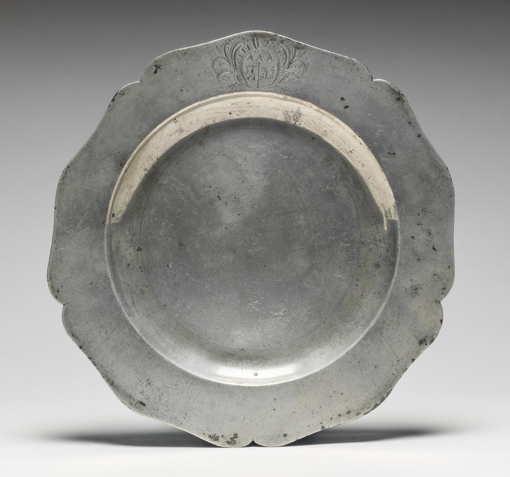 An image of Plate. Thomas Chamberlain of King Street, Soho. Pewter, cast and hammered, with engraved arms, height, whole, 2 cm, diameter, whole, 25 cm, circa 1750-1775. Circular with five indentations and five outward curves, curved sides and flat base. The underside of the rim has an applied raised edge. The rim is engraved with tan almost circular shield bearing he unregistered coat-of-arms of David Garrick (1717-79), surrounded and foliage. This plate was part of a service made for the celebrated actor and manager of the Drury Lane Theatre, David Garrick (1717-79). If the arms had tinctures (colours) they would be described: 'Per pale or and azure on the dexter compartment a tower gules and on the sinister on a mount vert a sea horse azure mane, fins and tail of the first, on a chief gold, three mullets of the second. (See Documentation, Burke) Thirty-one pieces from the service bearing either Garrick's arms or crest are at the Shakespeare Birthplace Trust at Stratford-on-Avon. A.F. de Navarro Bequest.