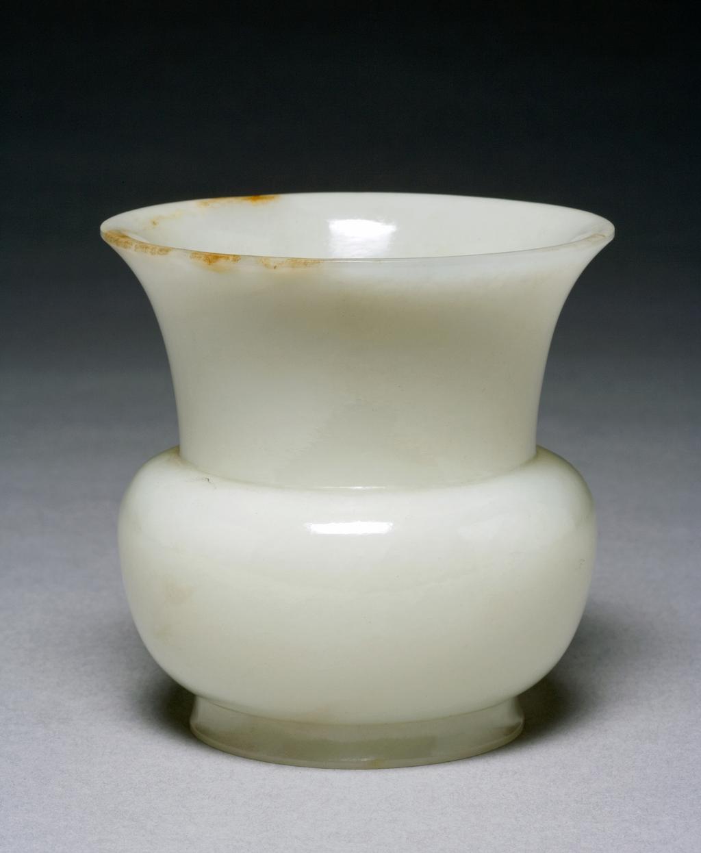 An image of Spittoon/Zhadou. White jade with brown areas on the rim, softly polish, well hollowed with the straight sides flaring out from the flat base before stepping sharply in at the shoulder and flaring out again to the wide trumpet mouth. Nephrite, height 8.5 cm, diameter, mouth, 8.5 cm, 1700-1800, Chinese.
