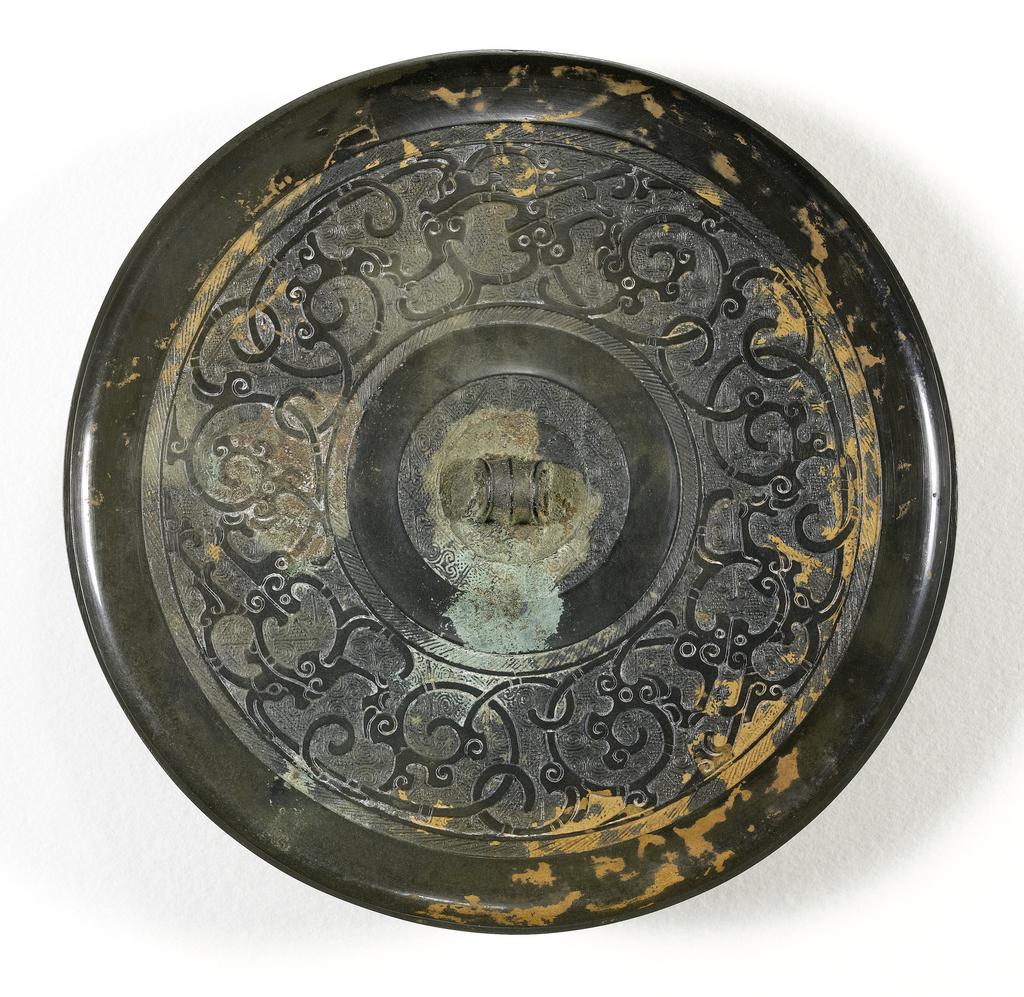 An image of Mirror made from bronze, of a glossy dark colour. Flanged edge, with mouth border. A wide band of much conventionalised dragons scrolled and intertwined, and a interlaced design on a diamond and spiral background. Inside is another smooth band, a hatched area, and the central boss-like loop for the cord. Bronze, diameter 18.3 cm. Chinese Period of the Warring States (481-221 BC).