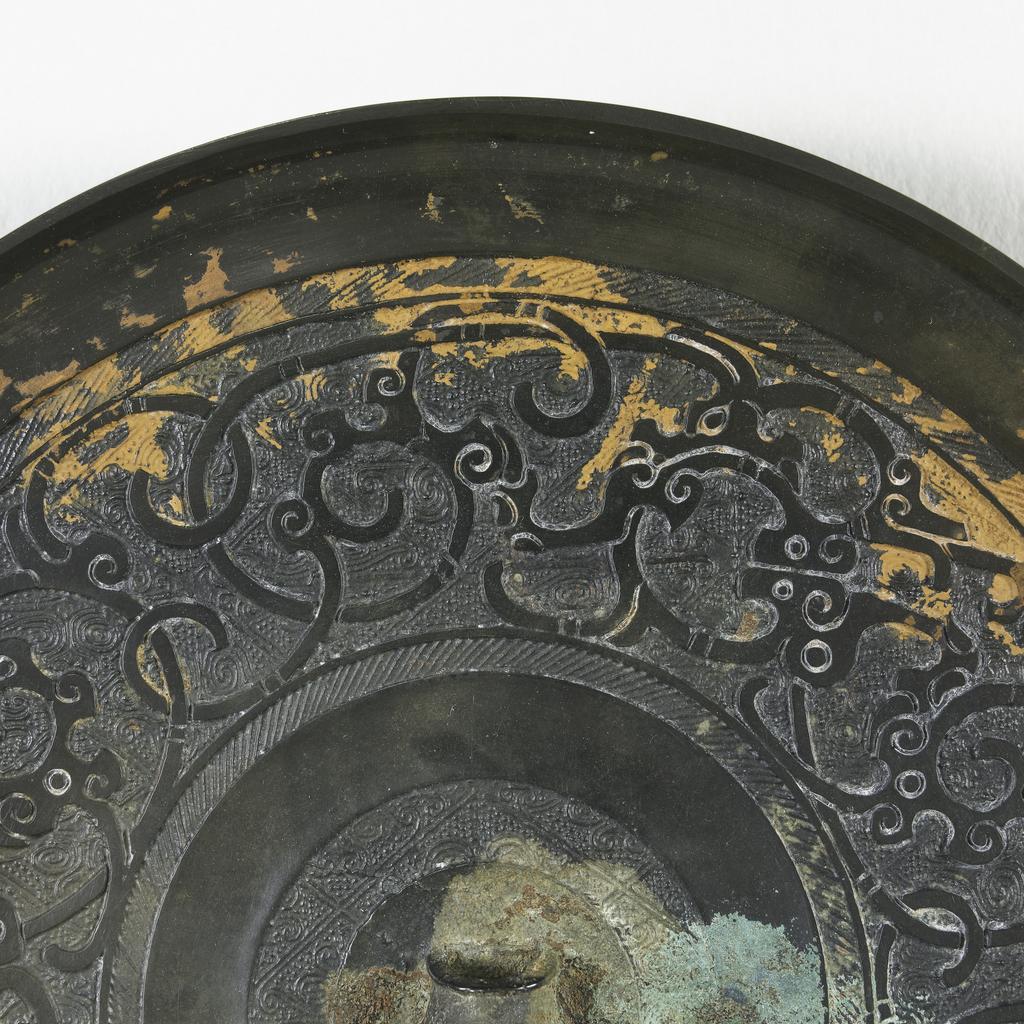 An image of Mirror made from bronze, of a glossy dark colour. Flanged edge, with mouth border. A wide band of much conventionalised dragons scrolled and intertwined, and a interlaced design on a diamond and spiral background. Inside is another smooth band, a hatched area, and the central boss-like loop for the cord. Bronze, diameter 18.3 cm. Chinese Period of the Warring States (481-221 BC).