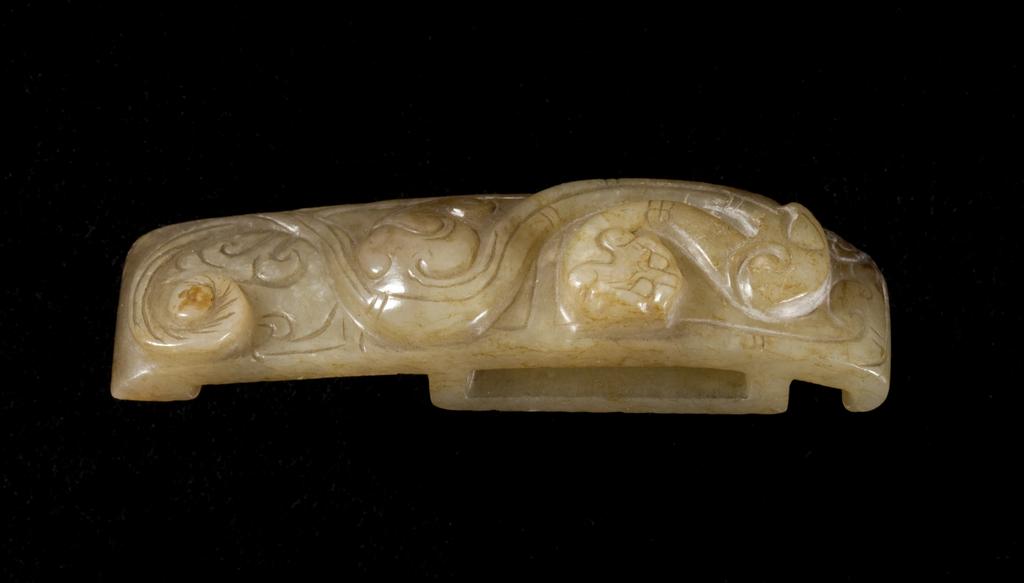 An image of Scabbard slide. Pale green translucent jade with some brown staining, decorated with a flattened version of a feline dragon. The underside of the piece is completely flat. Nephrite, height 1.25 cm, length 7.65 cm, width 1.9 cm, 1500-1700. Chinese.
