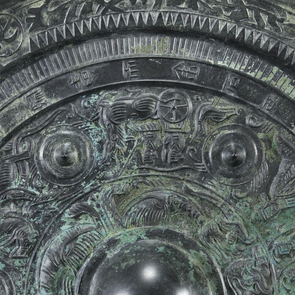 An image of Relief. Mirror made from bronze, inscribed with makers name (Teh'e). Around the central boss is a band of tail-less dragons, surmounted by a band of dragons, figures of men, and chariots. There is a band of inscription and a rim with dragons in light relief. Diameter 21.6 cm. Late Han Dynasty (206 BC-220 AD).