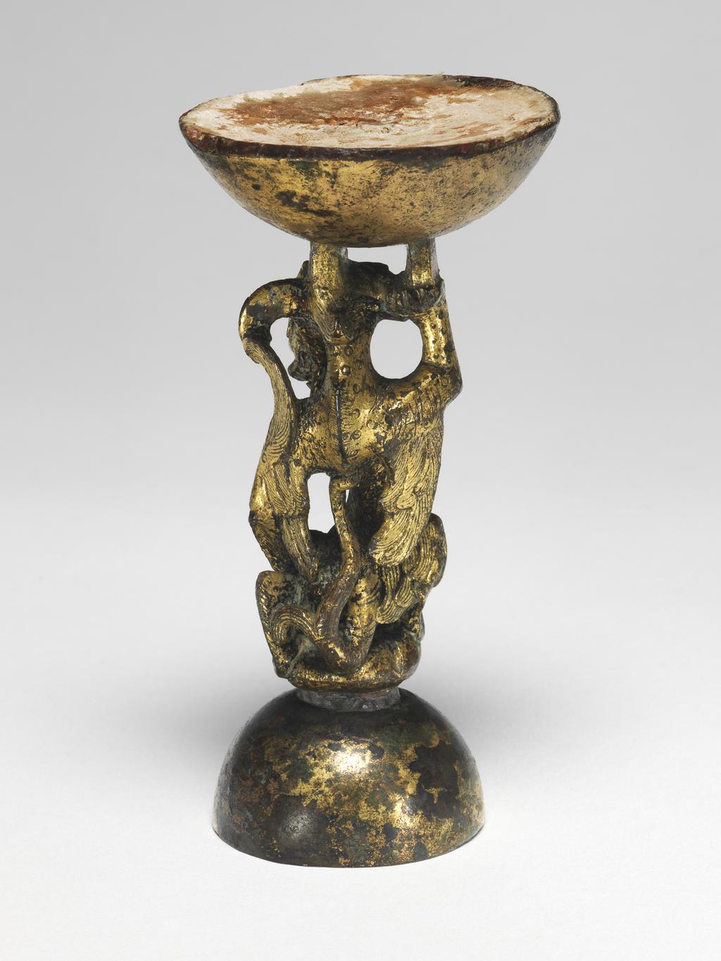 An image of Bronze stand in the shape of a dragon, rearing on its hind legs. The dragon is spotted, and kneeling on one knee with one paw on its hip. The other paw is supporting a lotus seed pod against its neck. Bronze, height 13.3 cm. T’ang Dynasty (618-906). Chinese.