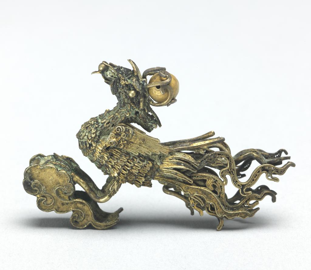 An image of Head of a hairpin. Hair ornament in gold filigree work. Unknown maker, China. It has the shape of a phoenix, which is standing on a cloud with extended tail feathers and with a raised head. The body and head of the phoenix are of thick plaited gold wires soldered together like feathers. The crest is in the form of a flower shaped receptacle for a gold bead. The phoenix is holding a bead in its beak. Beneath the cloud is a hole for fixing the bird to a head dress. Gold filigree, soldered, width 4.2 cm, height 3.8 cm. Tang Dynasty (618-907).