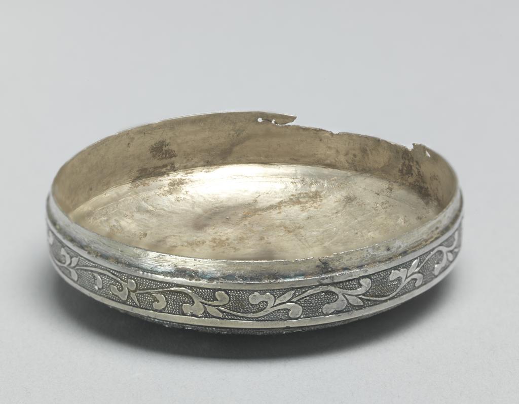 An image of Box. Unknown maker, China. Small circular box, made from beaten silver, of a slightly convex shape with straight sides. Both the box and the cover are decorated with foliate scrolls. The pattern has been gilded and is chased on a ground of regular ring matting. The sides have been soldered to the base. Silver, chased, gilded and engraved, diameter 5.2 cm, height 1.9 cm, T’ang Dynasty (618-906).